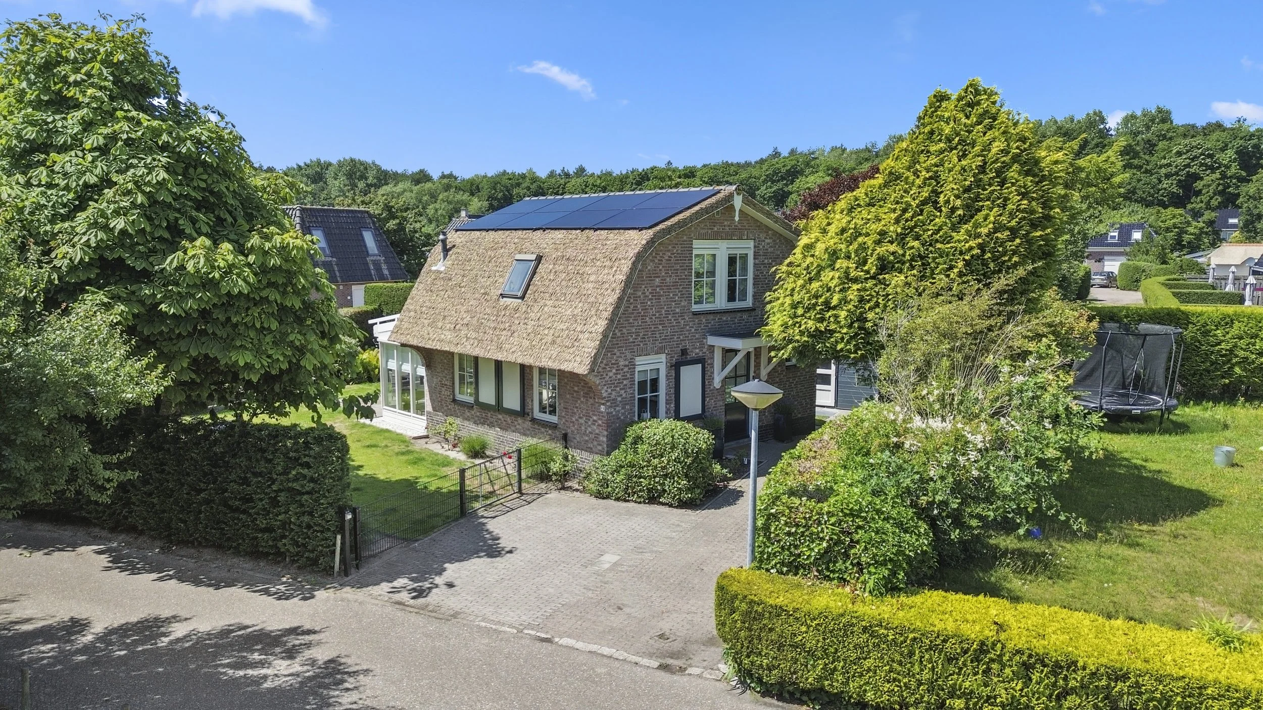Gezicht op een huis met een rieten dak en zonnepanelen, omgeven door groene bomen en een tuin, onder een blauwe lucht.