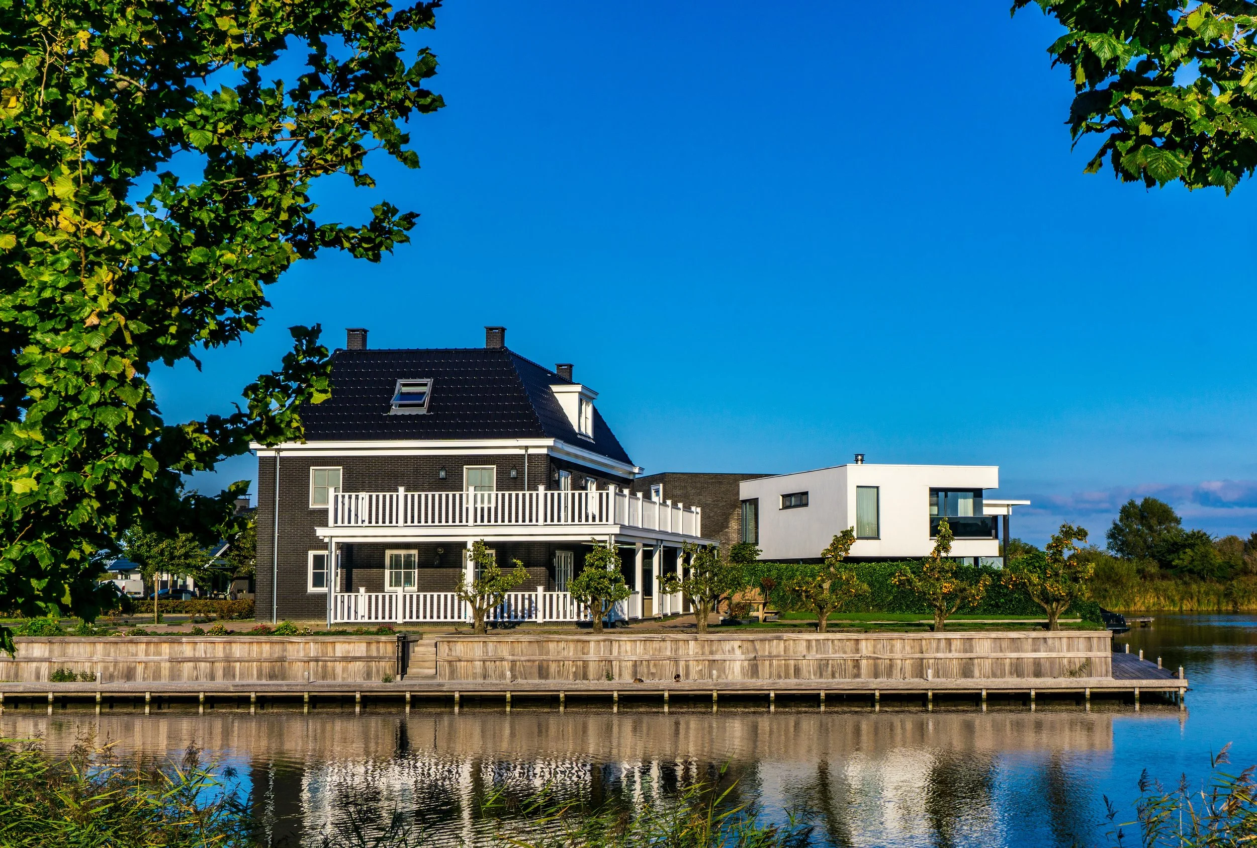 Moderne huis aan een rivier met een houten wal, omgeven door groene bomen en een heldere blauwe lucht.