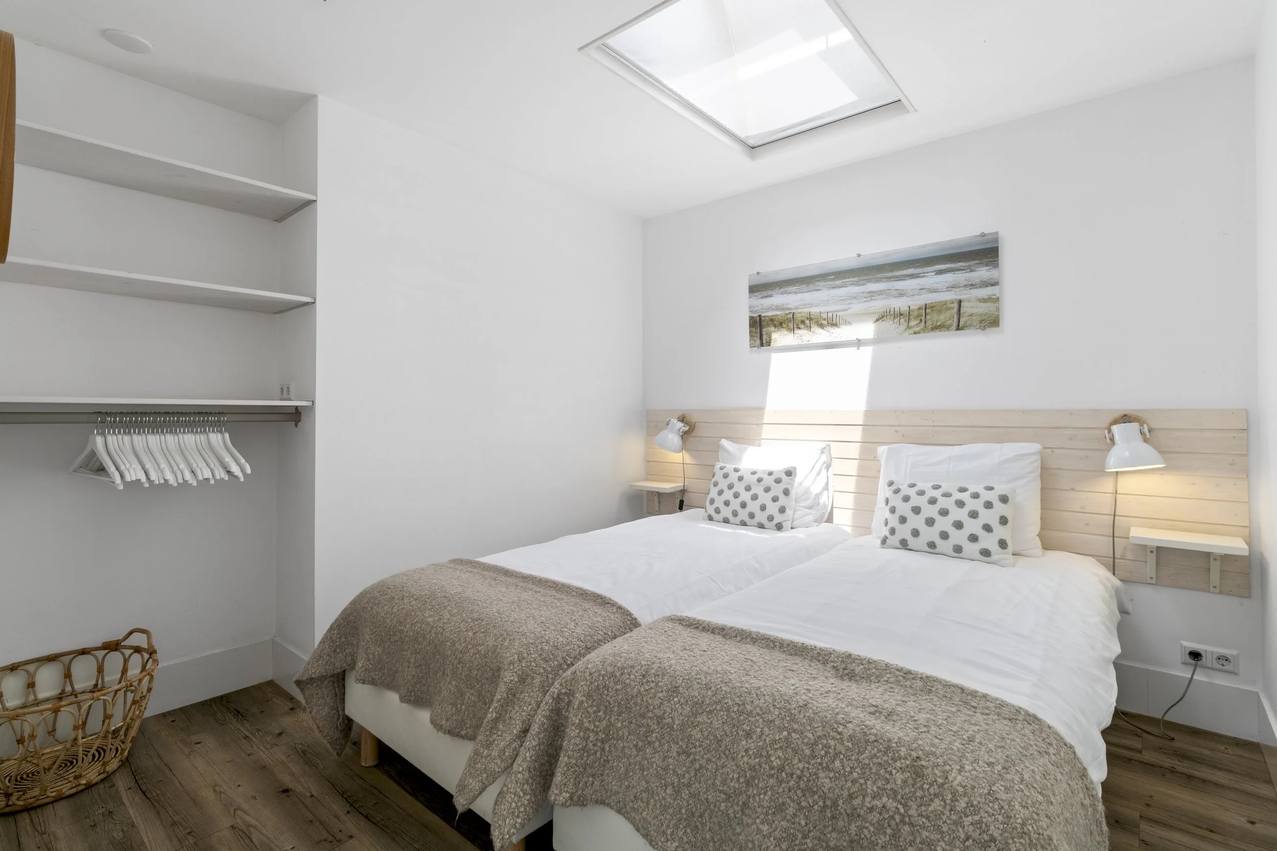 Een witte slaapkamer met twee eenpersoonsbedden met beige dekens, kussens met stippen, wandlampen en een raam in het plafond.