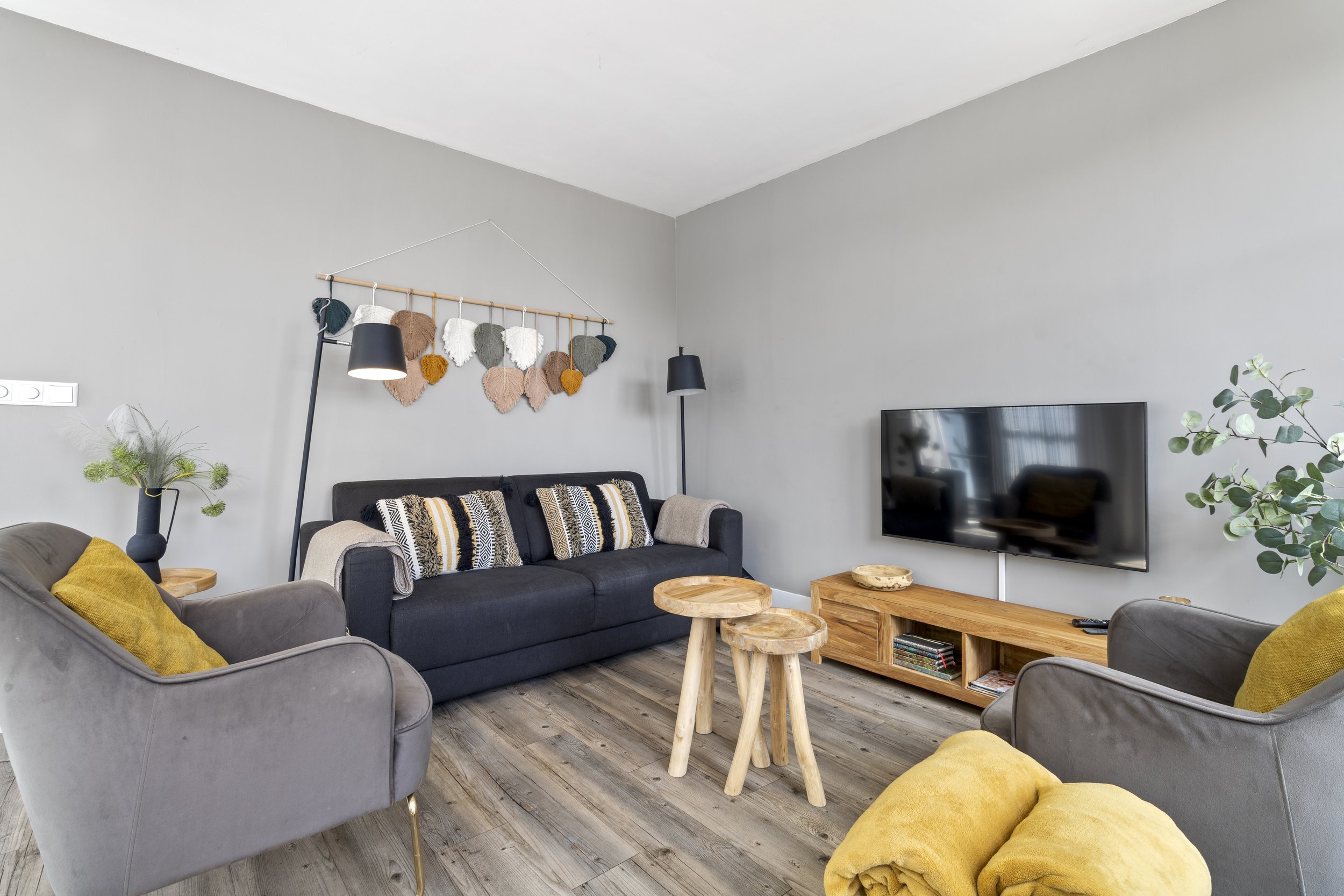 Woonkamer met grijze en gele fauteuils, houten salontafels, zwart bank, plant, tv, muurdecoratie en houten vloer.