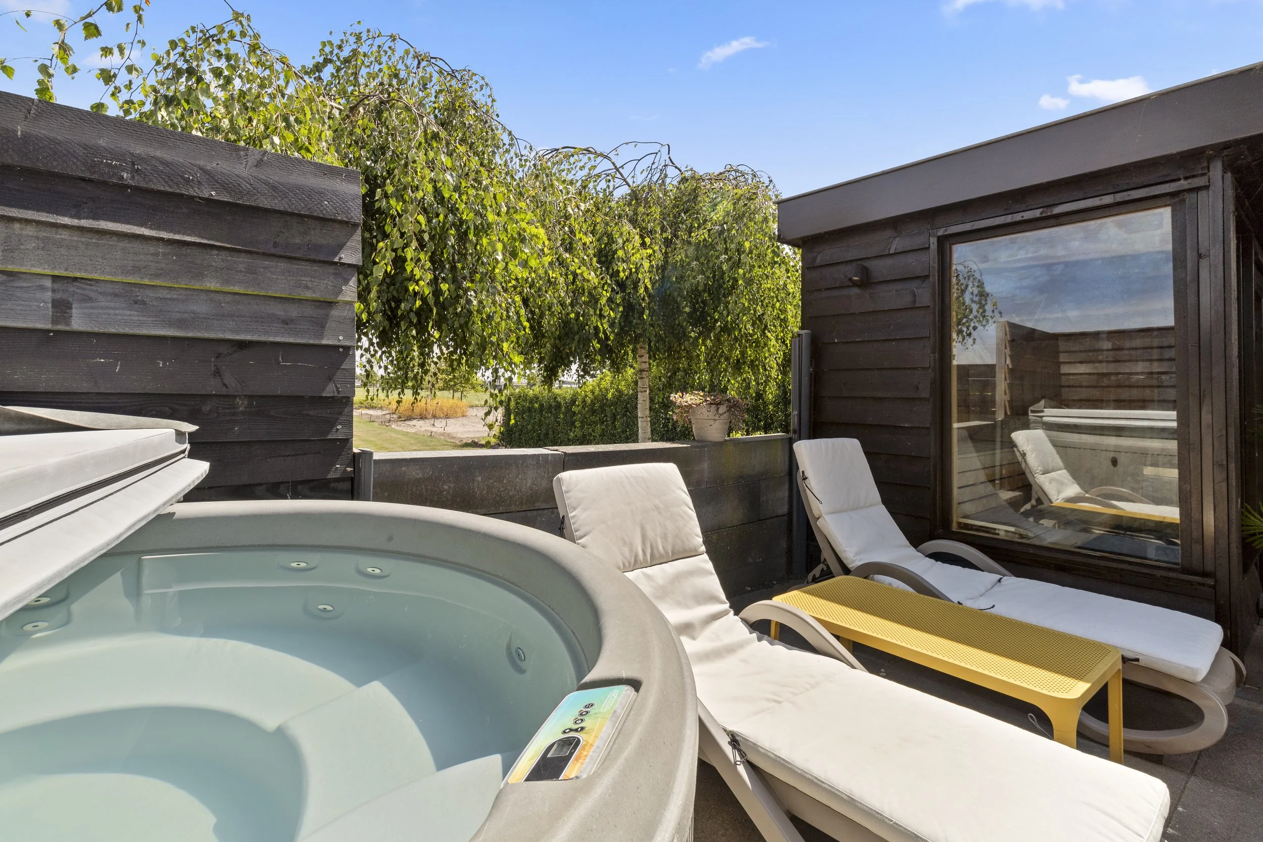 Buitenruimte met ligbedden en een jacuzzi, omgeven door groene bomen onder een blauwe lucht
