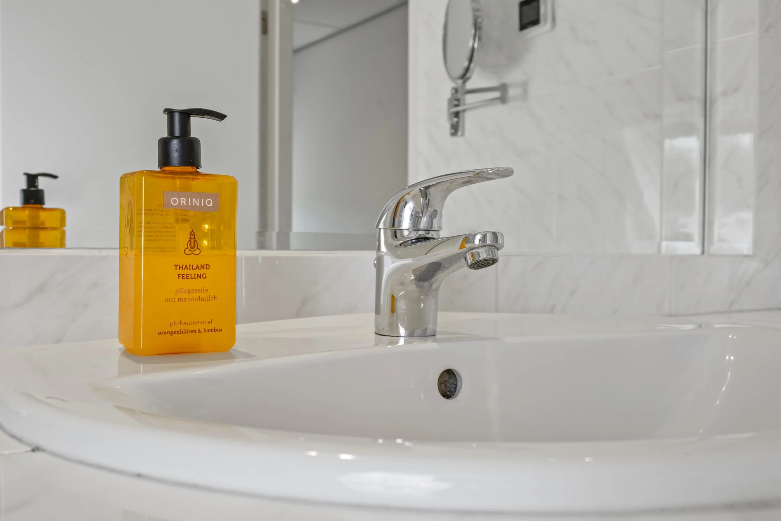 Een witte badkamer wastafel met een zilveren kraan en een flesje zeep met oranje kleur, naast een spiegel