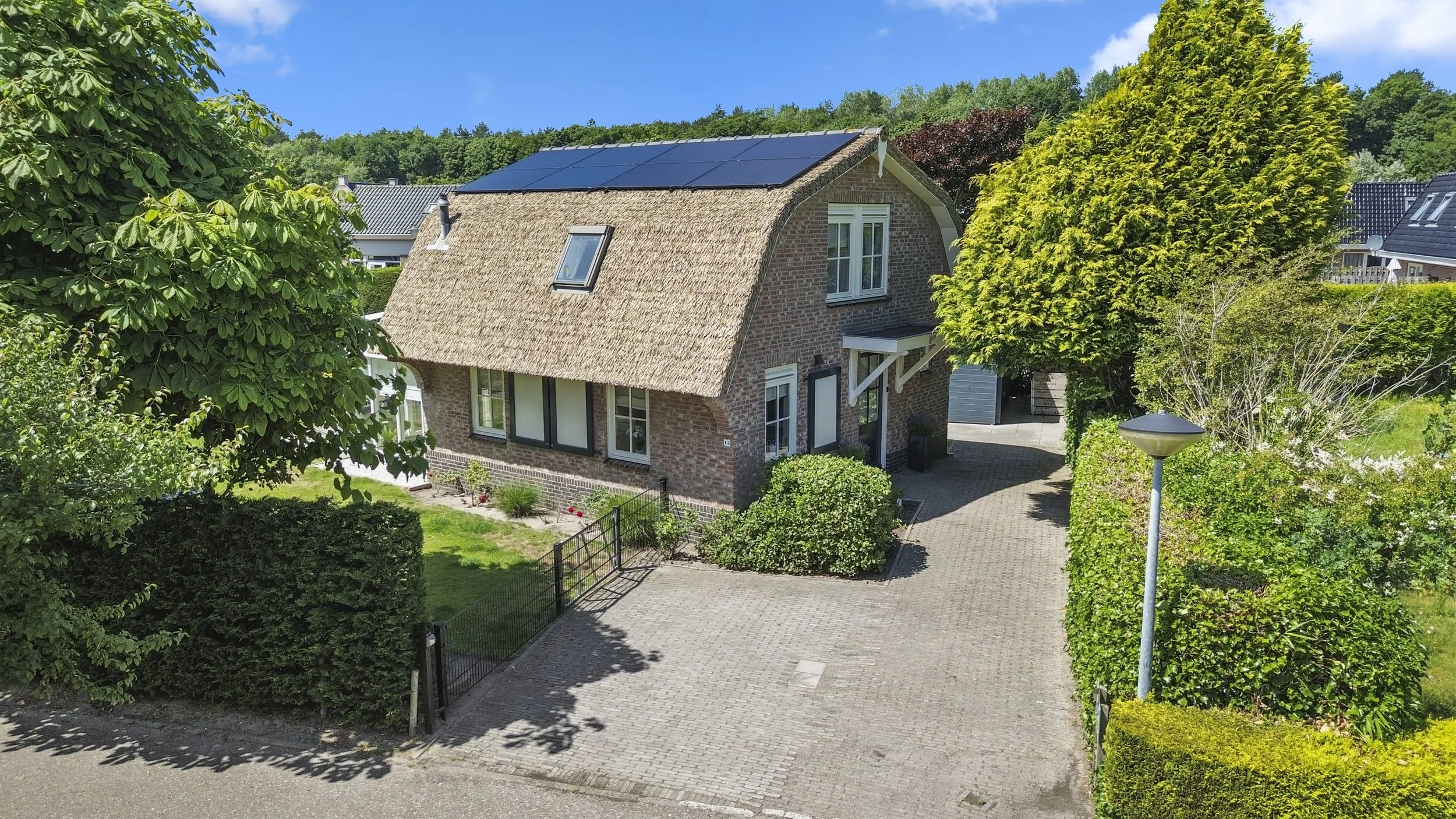 Een huis met een rieten dak, zonnepanelen, omringd door groene bomen en heggen, met een oprit en een tuin onder een blauwe hemel.