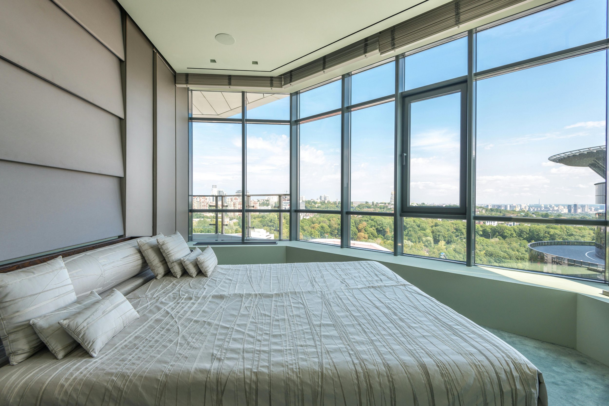 Een slaapkamer met een groot bed en uitzicht over de stad en groene parken via grote ramen.