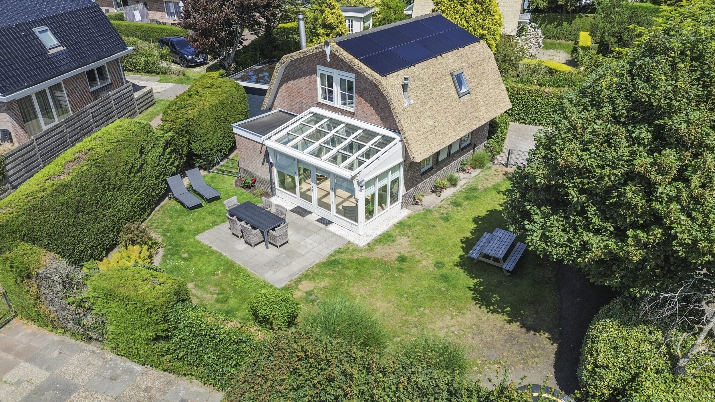Luchtfoto van een tuin met een huis dat een serre en zonnepanelen op het dak heeft, meerdere bomen, zitgedeeltes, en een omheinde tuin, in een groene en rustige buurt.