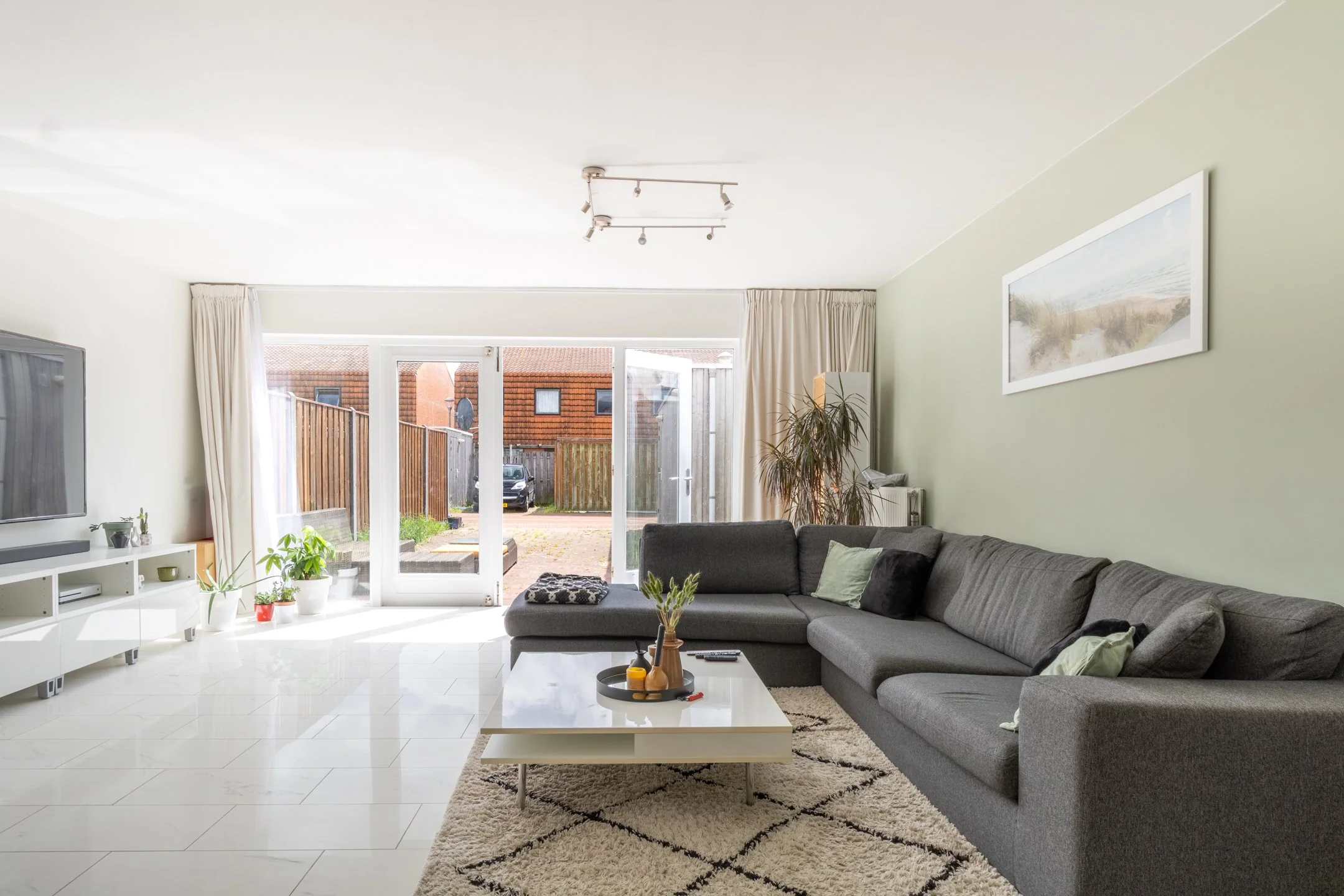 Modern woonkamer met grote hoekbank, tv, planten en uitzicht naar tuin met open deur.