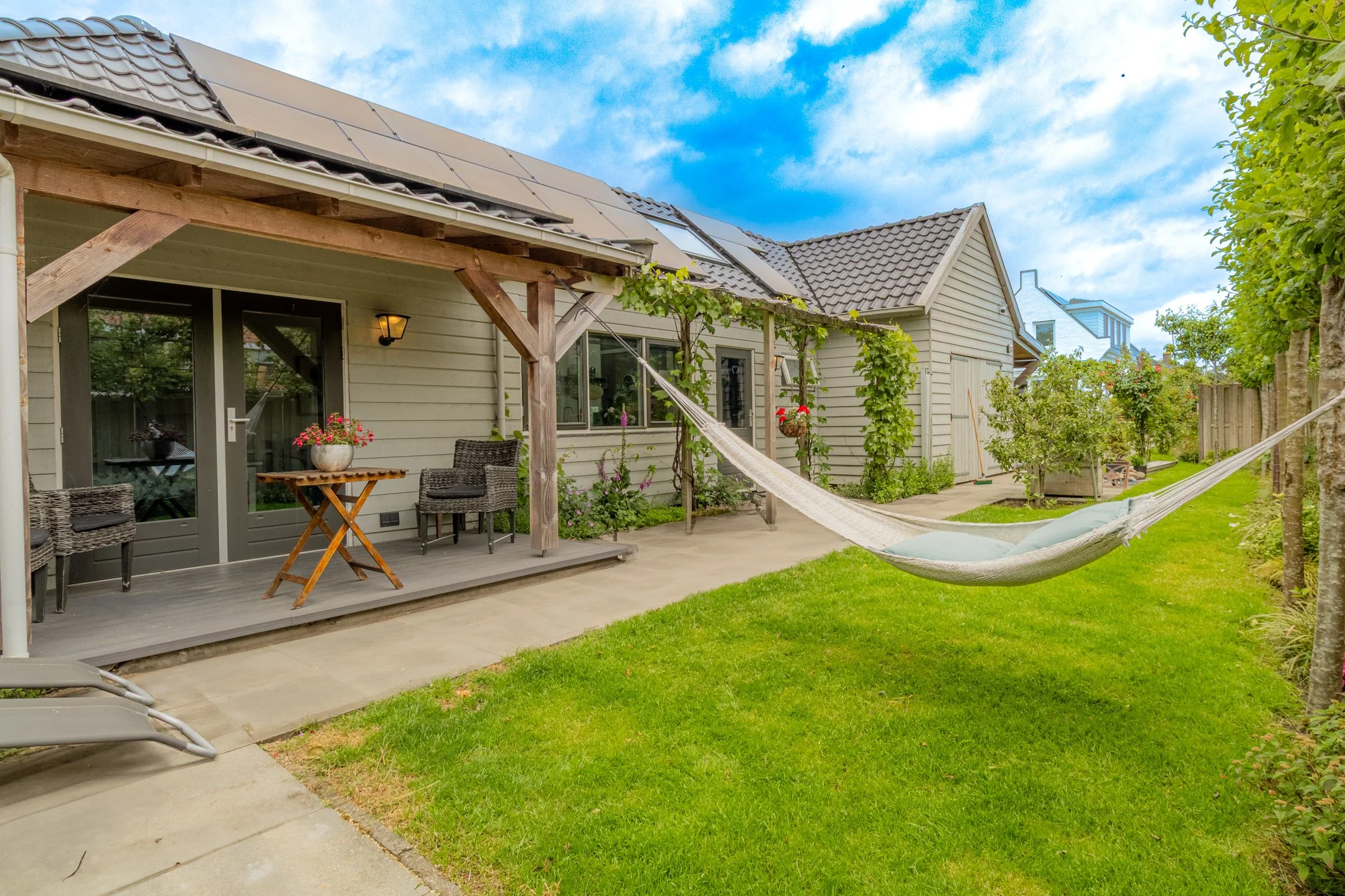 A backyard with a patio, a hammock, green grass, and plants, adjacent to a house with solar panels on the roof.