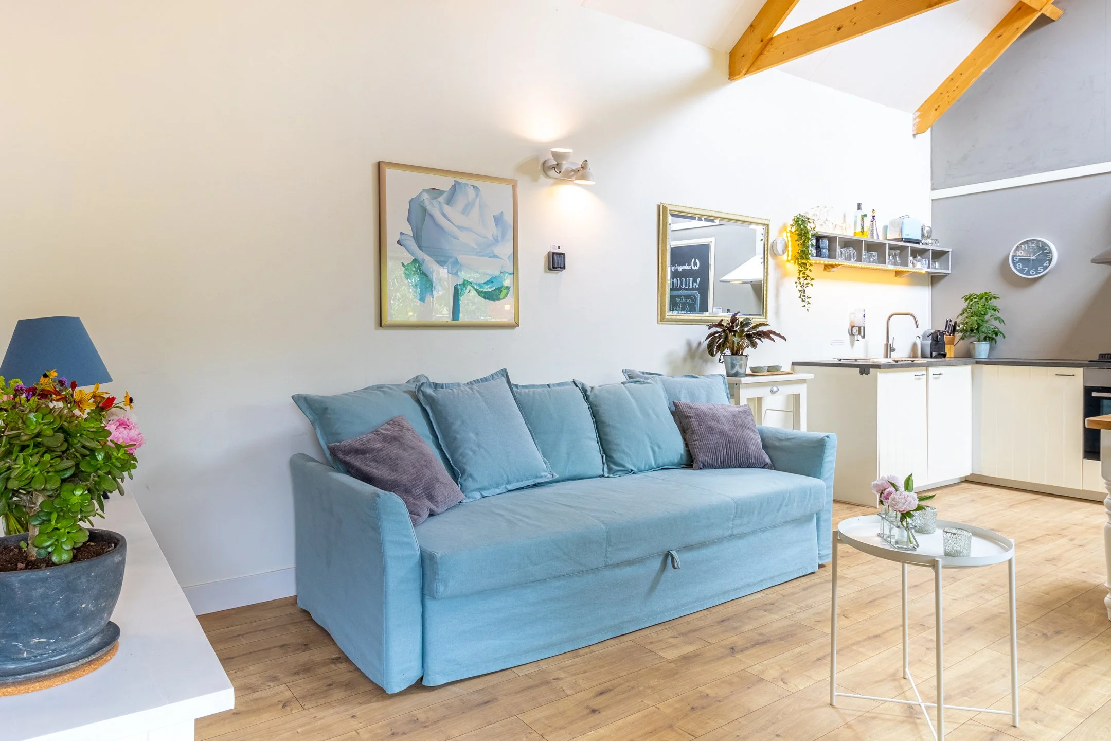 Woonkamer met een lichtblauw bank, een witte tafel met bloemen, een boekenkast met wijnflessen, en een kitchenette in een ruimte met een hoge plafond en houten balken.