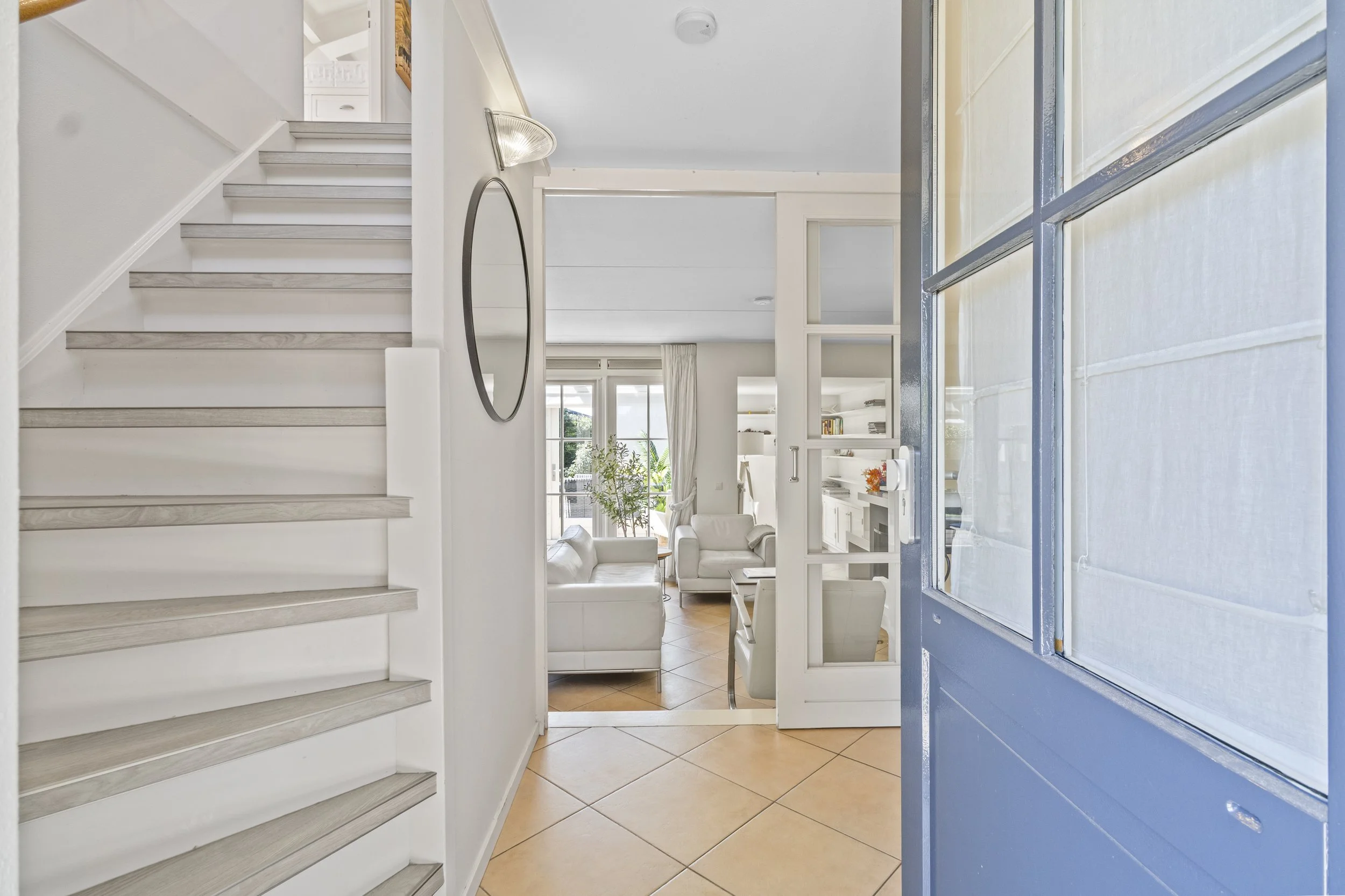 Binnenkijkje in een wit ingericht woonkamer via de entree met een blauwe voordeur, trap en ronde spiegel aan de muur.