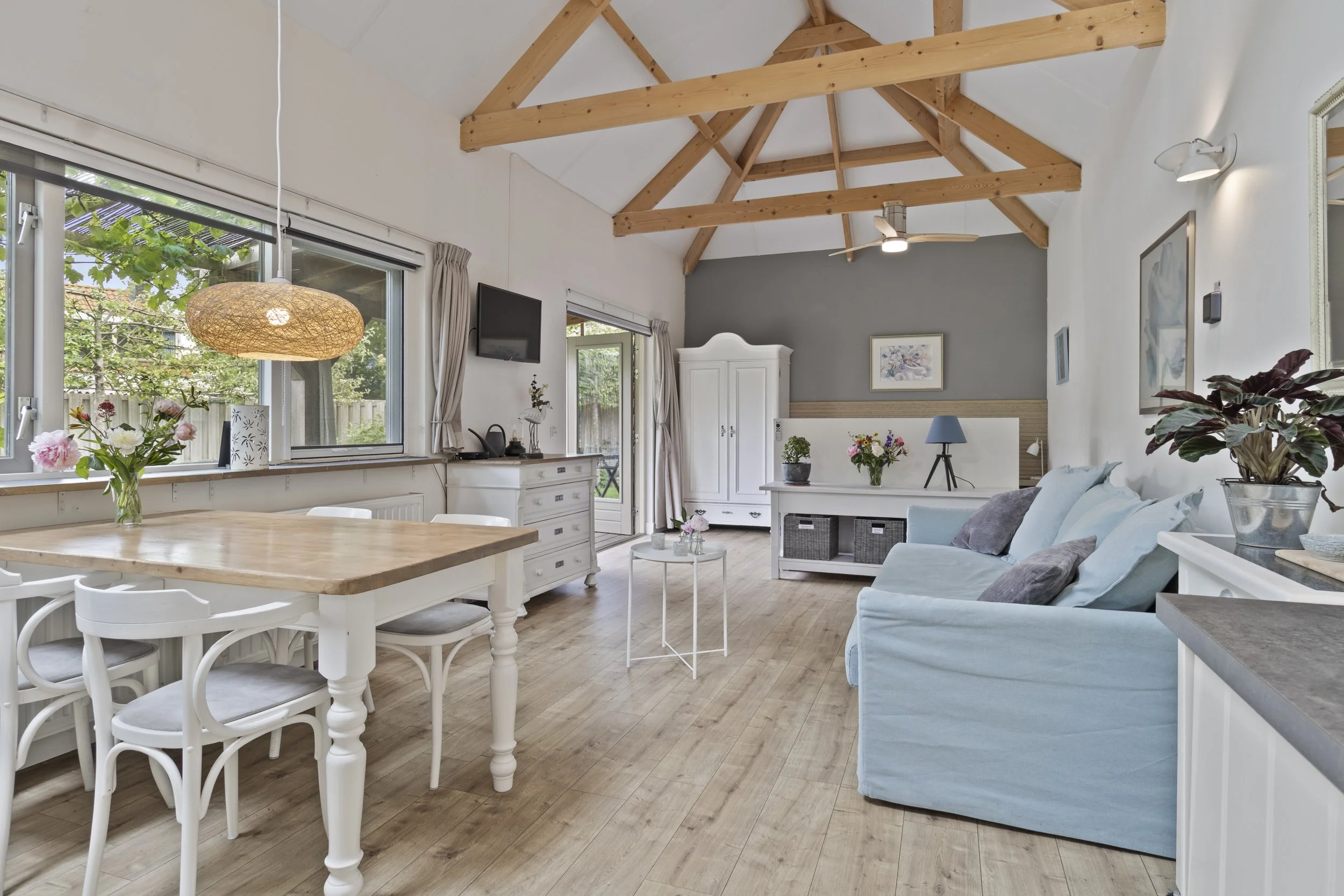 Lichte woonkamer met houten vloer, witte muren, een lichtblauwe bank, witte kasten, een eetkamertafel met witte stoelen en grote ramen met uitzicht op de tuin.