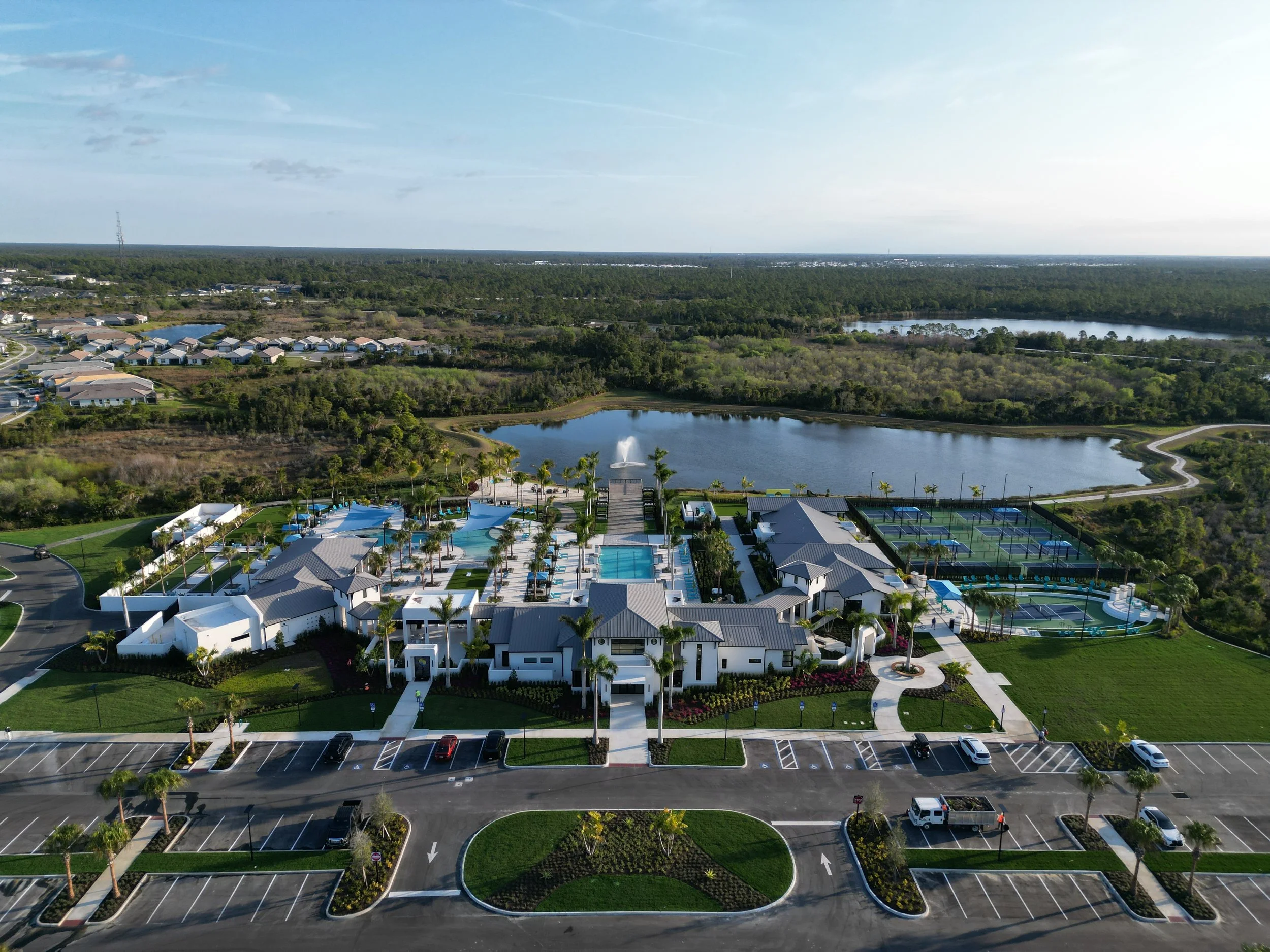 Aerial view of a residential area with houses, a pond, and a community pool area, surrounded by green trees and open land.