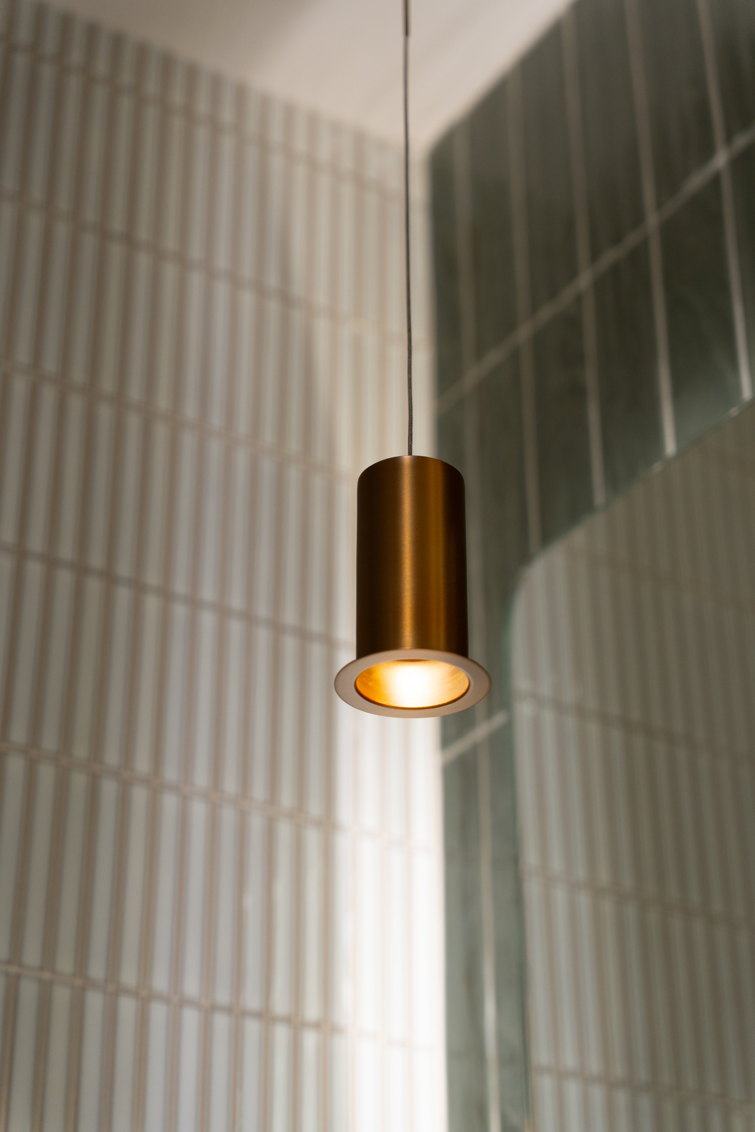 A modern pendant light fixture hanging from a ceiling, with a reflection and contrasting textured wall surfaces in the background.