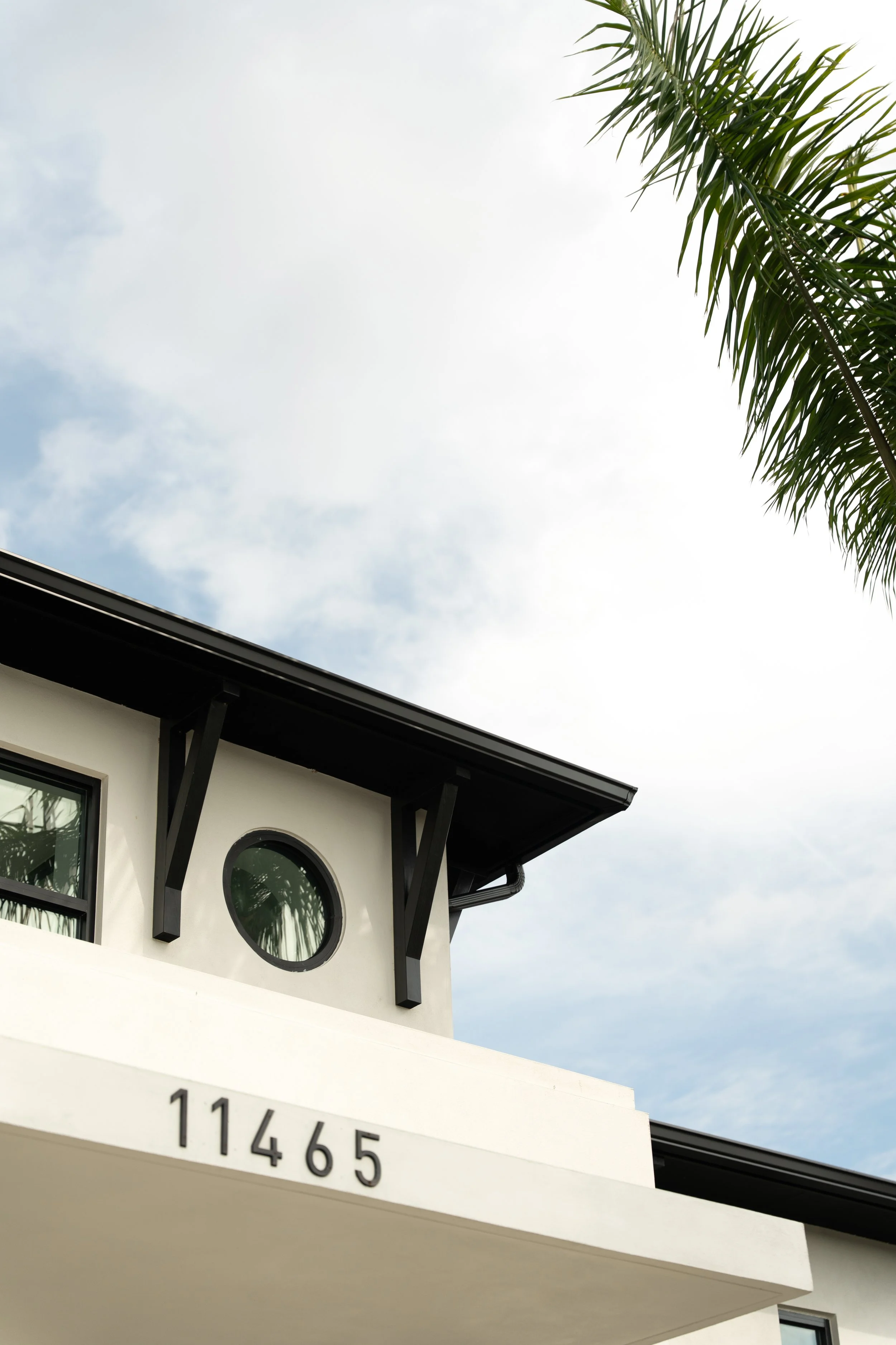 Part of a modern house with white exterior walls, round and rectangular windows, and a black roof edge. There is a palm tree in the background, and the house number 11465 is visible on the front.