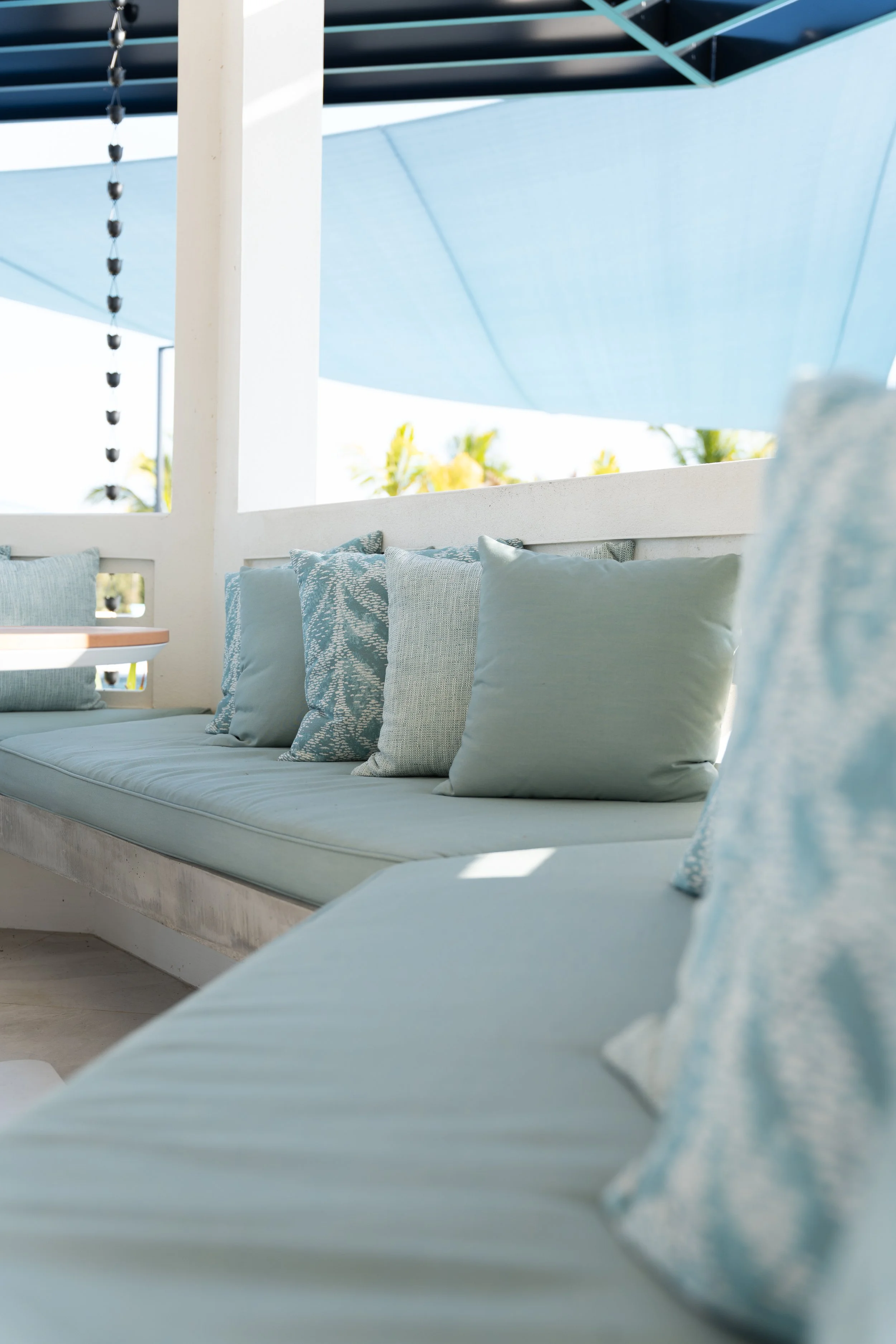 A row of light blue and patterned throw pillows on a cushioned outdoor bench under a blue shade sail, with a modern white and blue patio setting.
