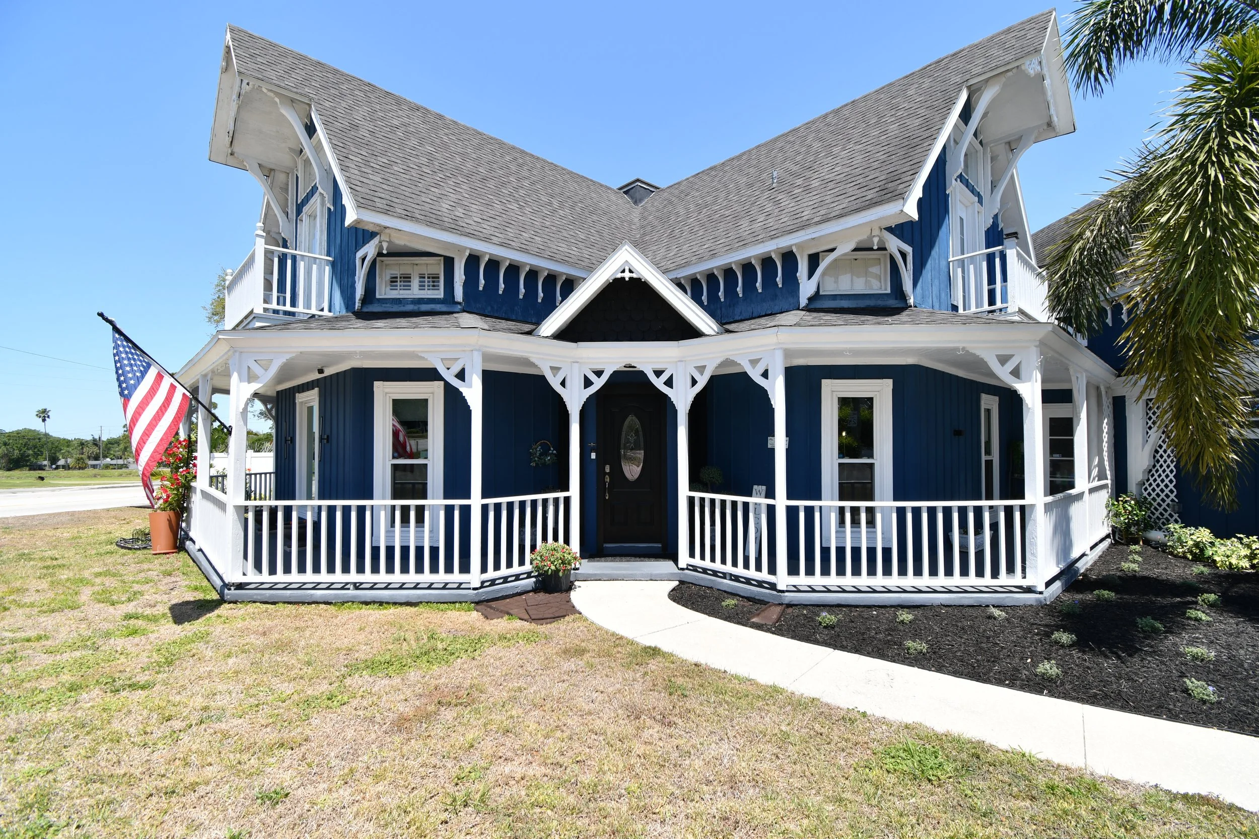 A two-story house painted blue with white trim and decorative woodwork. It has a wraparound porch with a white picket fence, potted plants, and an American flag. The roof is steeply pitched with dormer windows, and the sky is clear and blue.