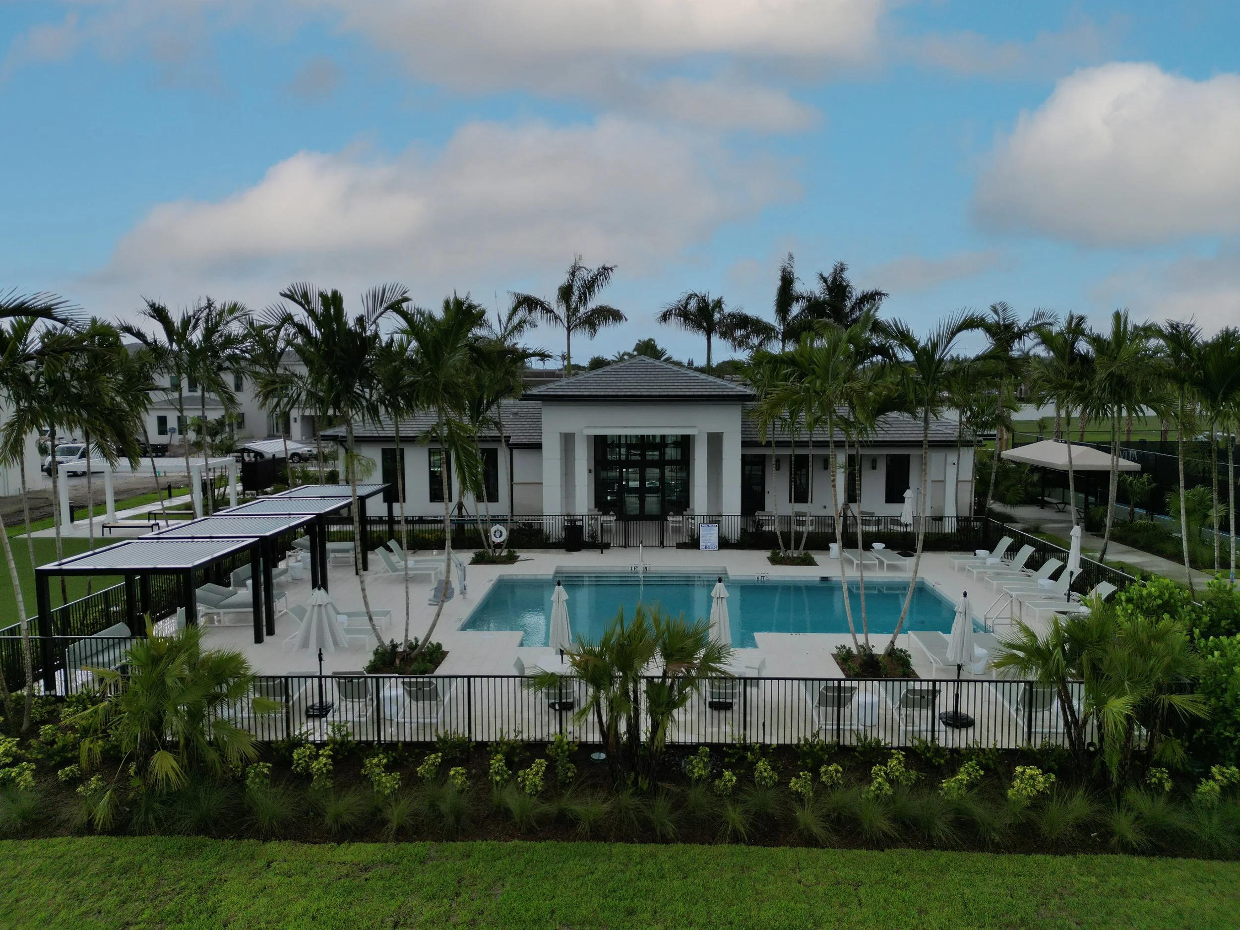 A residential backyard with a swimming pool, surrounded by lounge chairs and umbrellas, tall palm trees, and a modern house with a gray roof in the background. The sky is partly cloudy with blue patches.