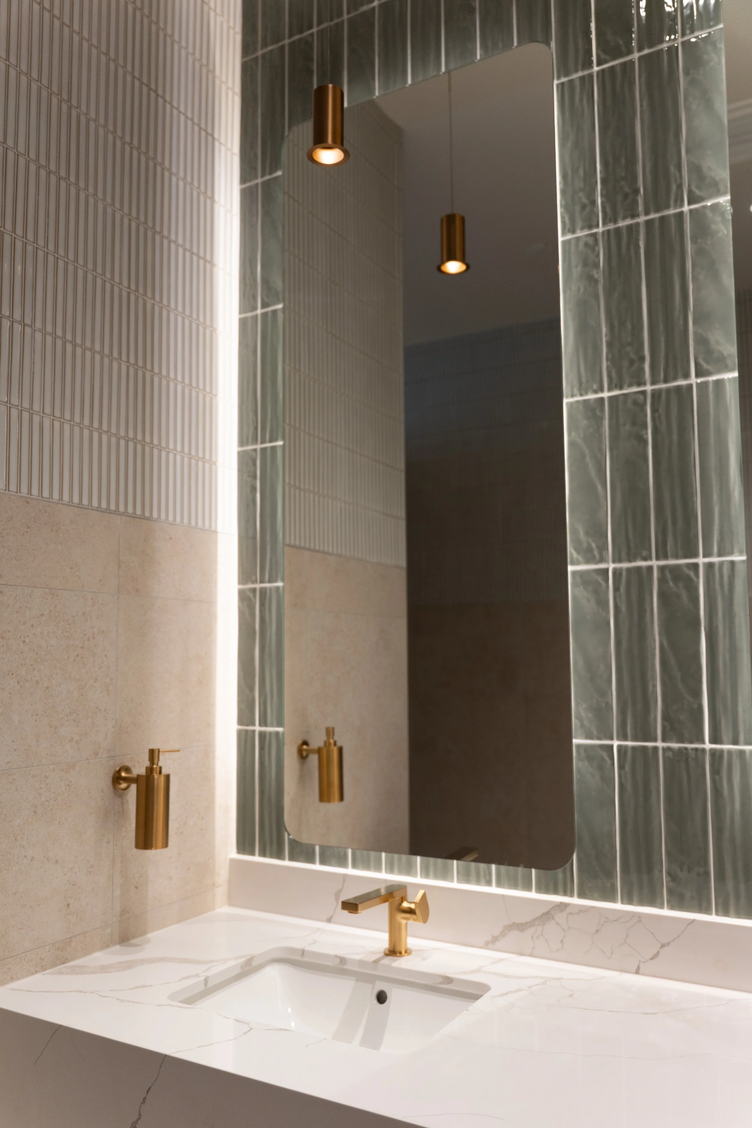 Modern bathroom with a large mirror, green marble wall tiles, gold fixtures, and a white marble countertop with a sink.