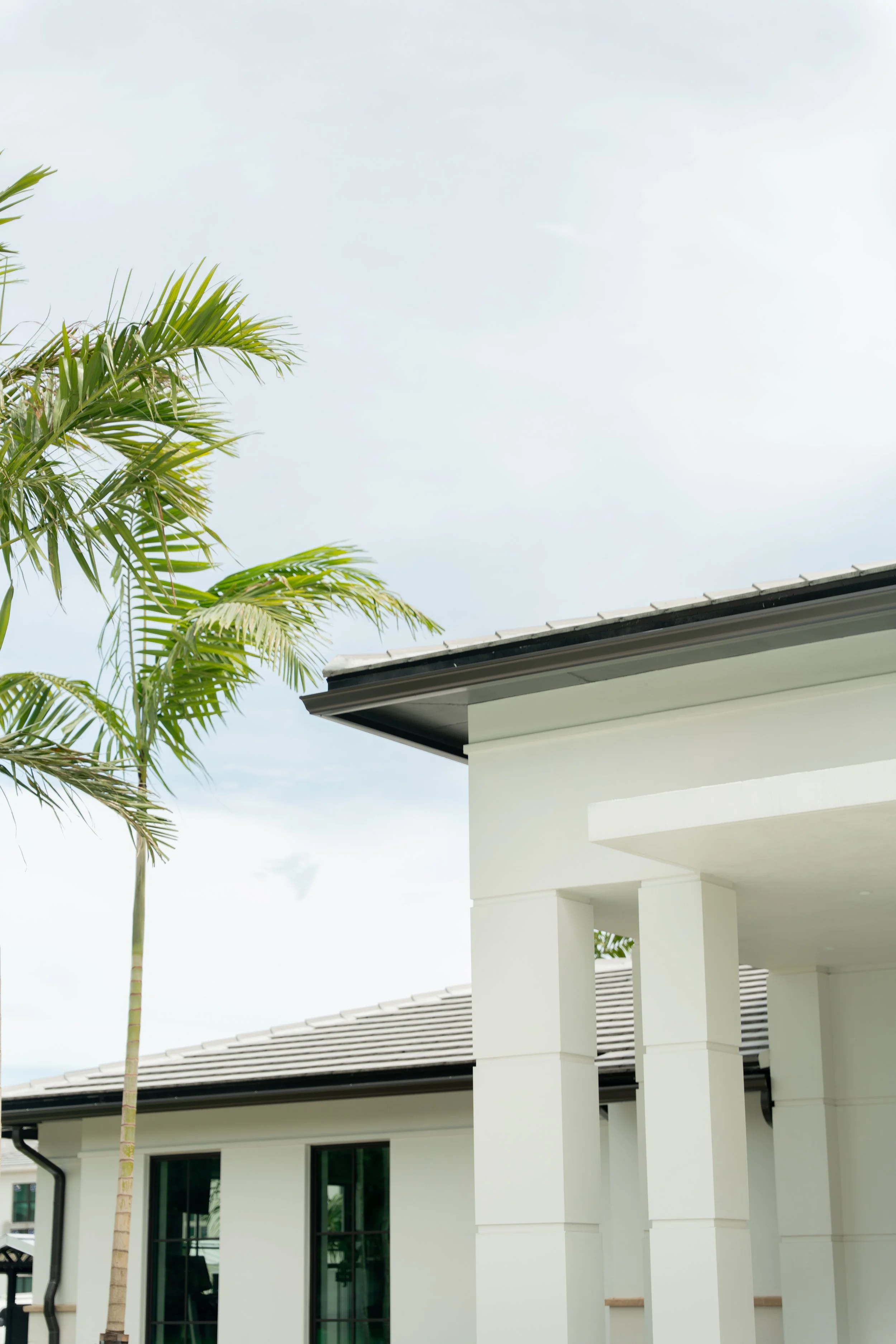 Part of a modern white house with large windows, white columns, and a roof with black edges. A tall palm tree is on the left side of the image, and the sky is partly cloudy.