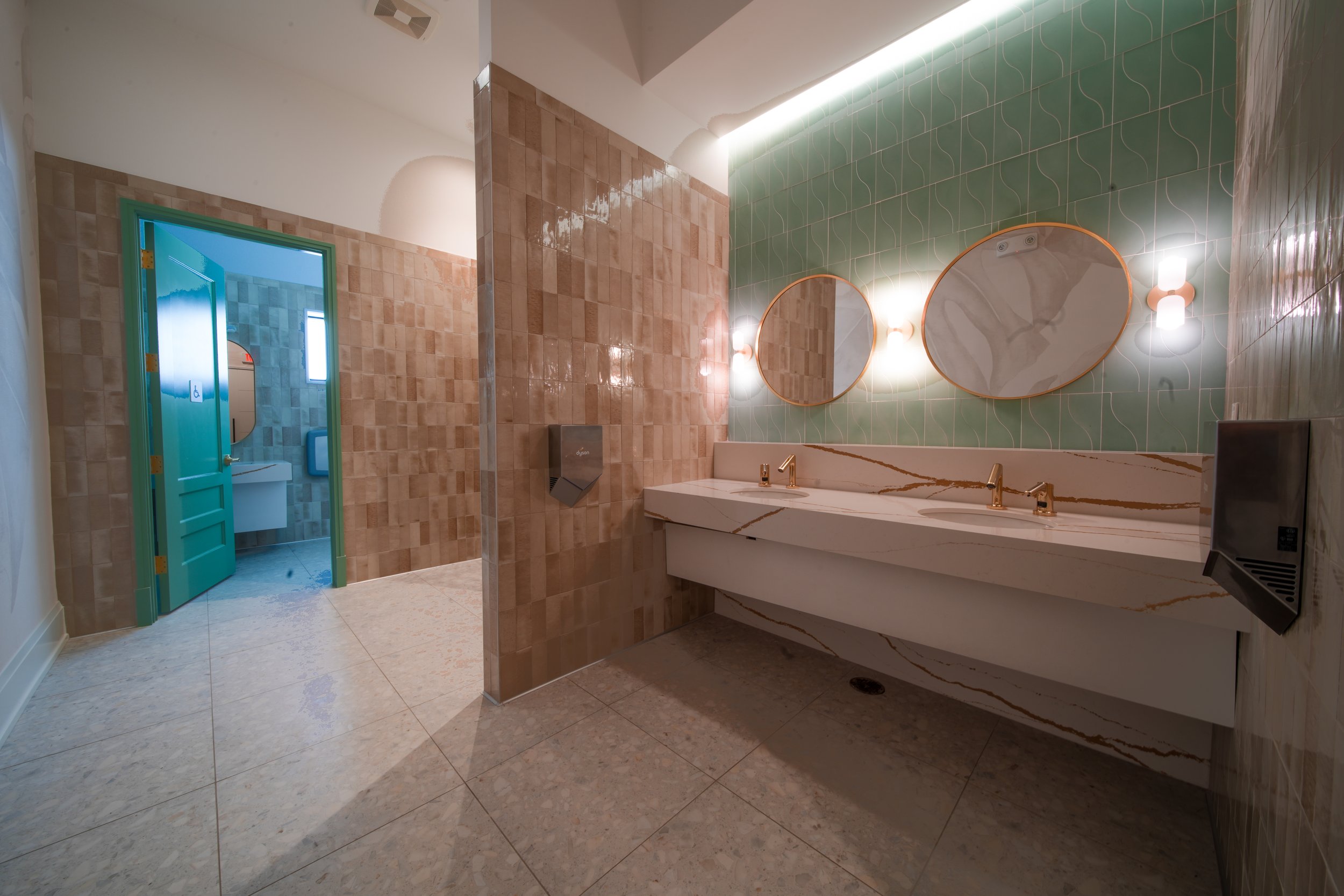 A modern restroom with dual sinks, round mirrors, green and peach wall tiles, and a door leading to a handicap accessible bathroom.