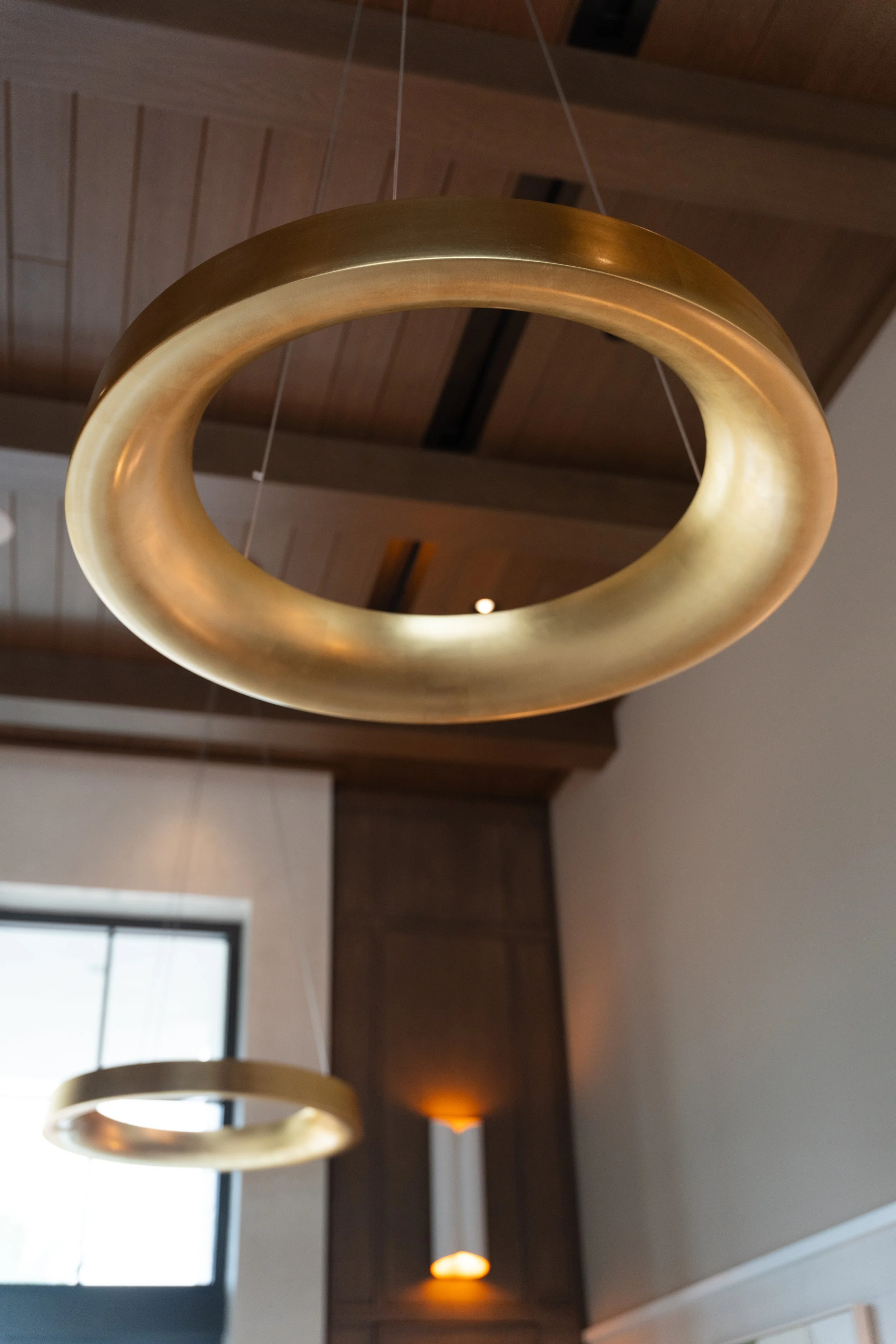 Close-up of a modern, oval-shaped, metallic gold pendant light hanging from a ceiling with visible wires, with a wooden ceiling and windows in the background.