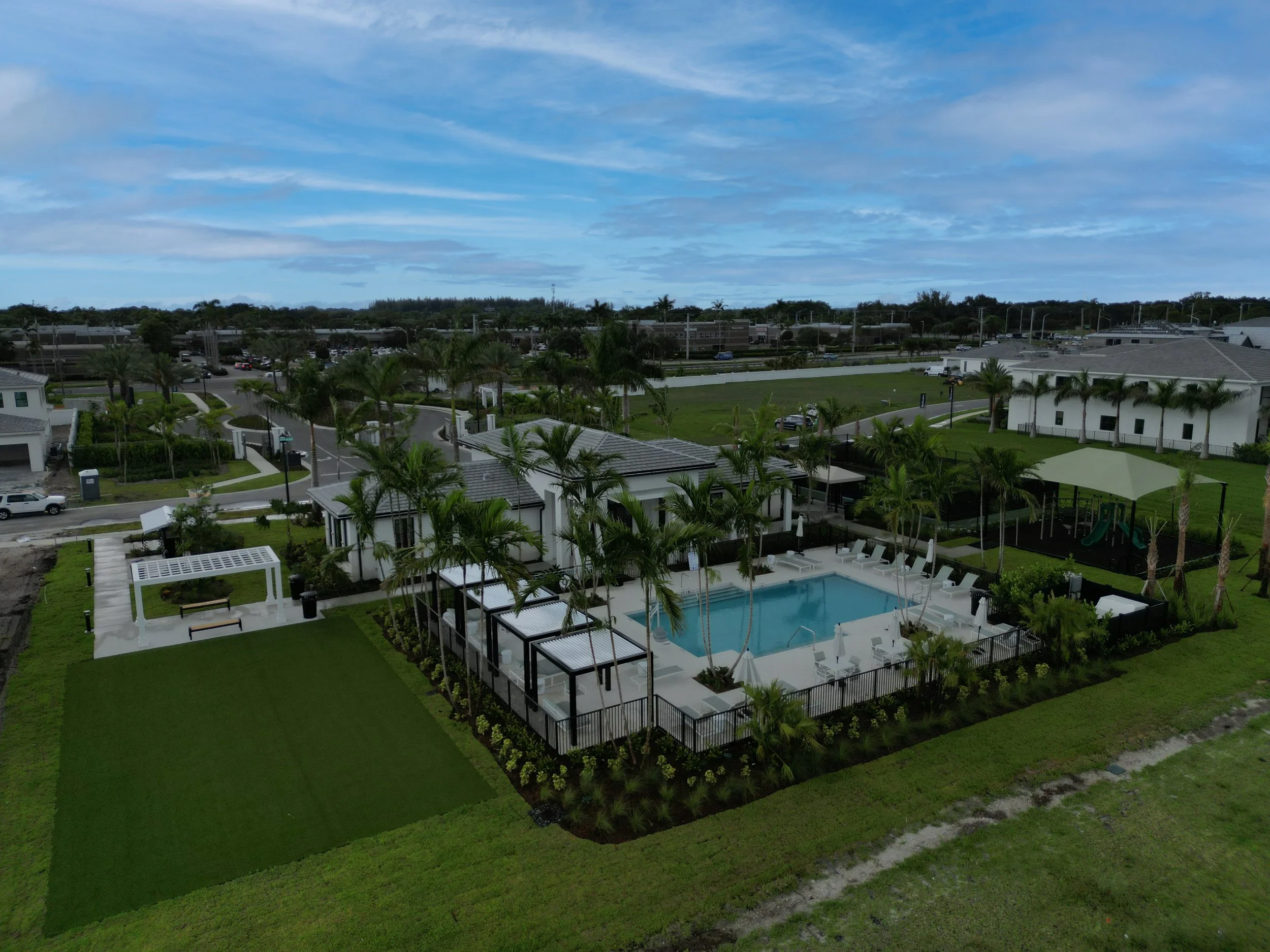 A residential area with white houses, palm trees, a swimming pool, and landscaped lawns, viewed under a partly cloudy blue sky.