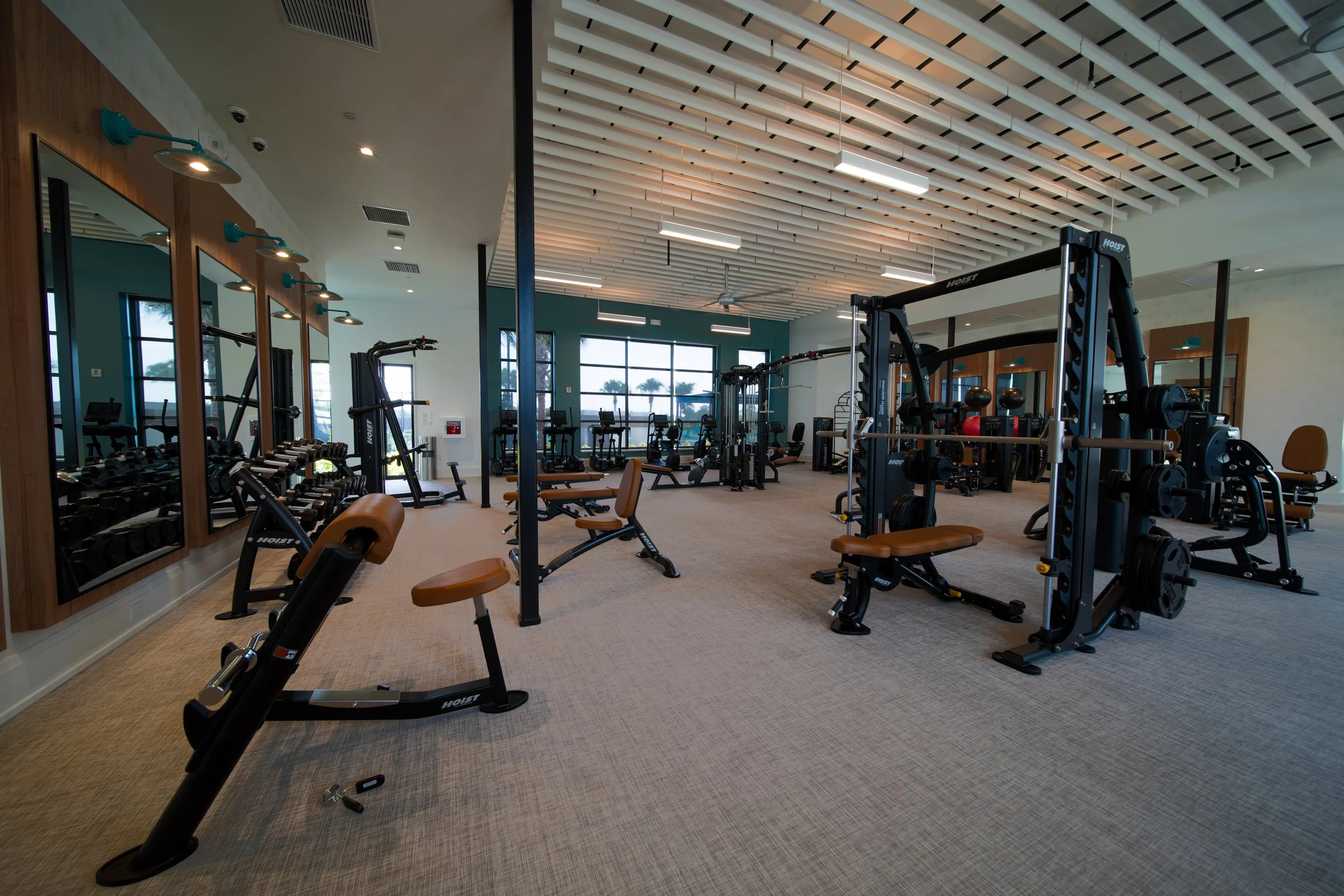 A modern gym with various workout equipment, including weight benches, dumbbells, and cardio machines, lit by overhead lighting and large windows.