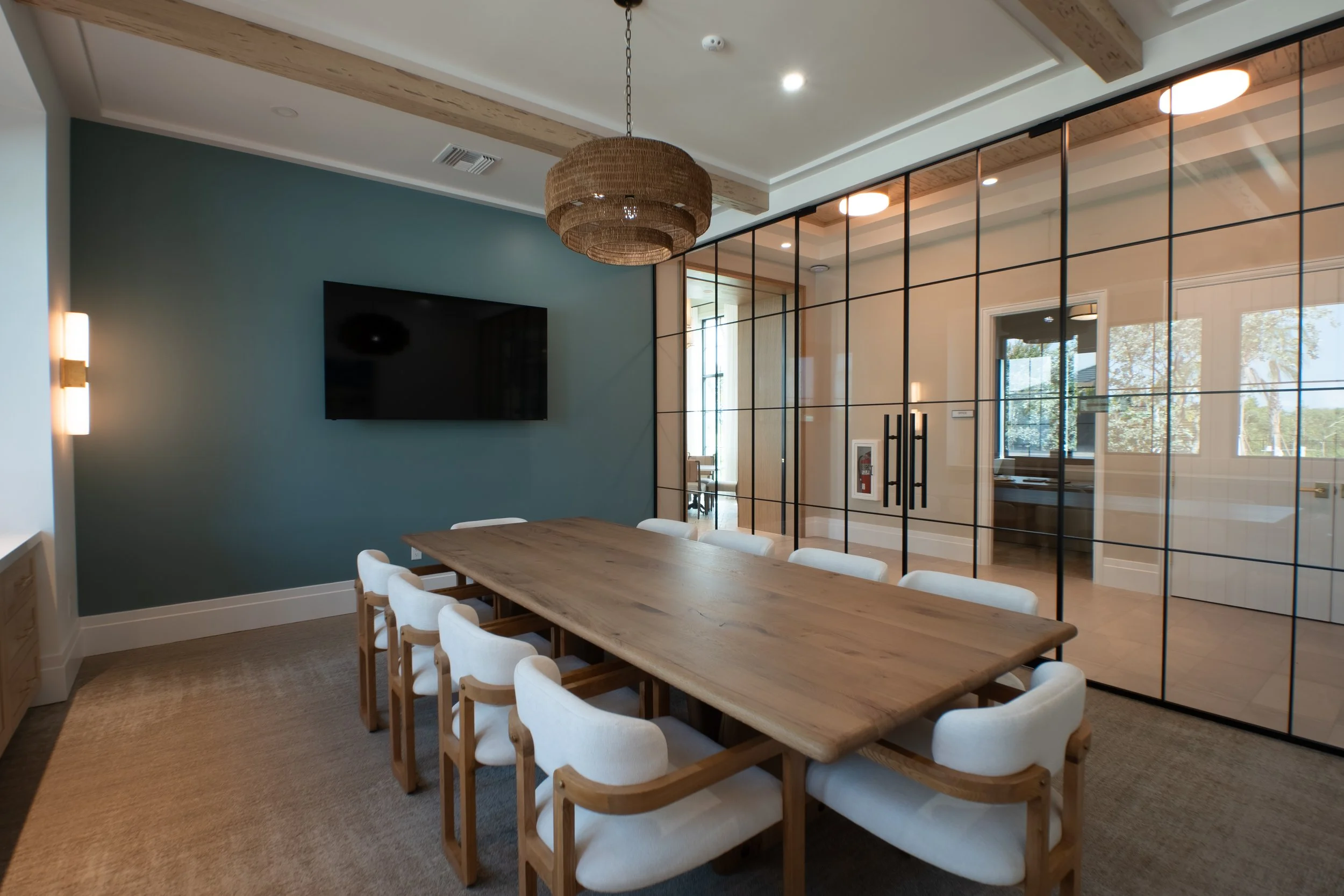 A modern dining area with a large wooden table surrounded by white upholstered chairs, a green accent wall with a mounted flat-screen TV, and a ceiling with wood beams and a woven light fixture, separated from another room by a glass partition with b