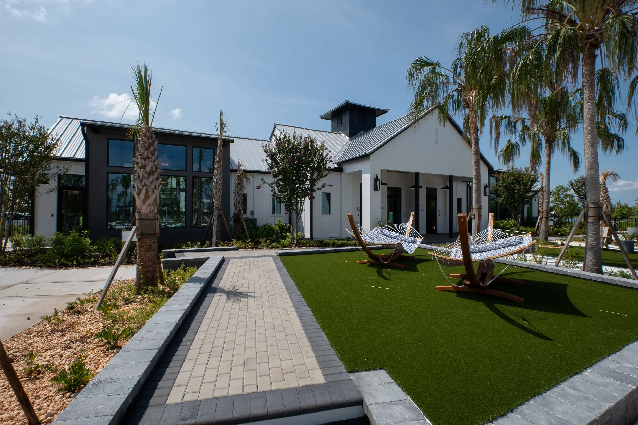 Modern house with large glass windows, metal roof, surrounded by palm trees, hammock, and paved walkway on a sunny day.