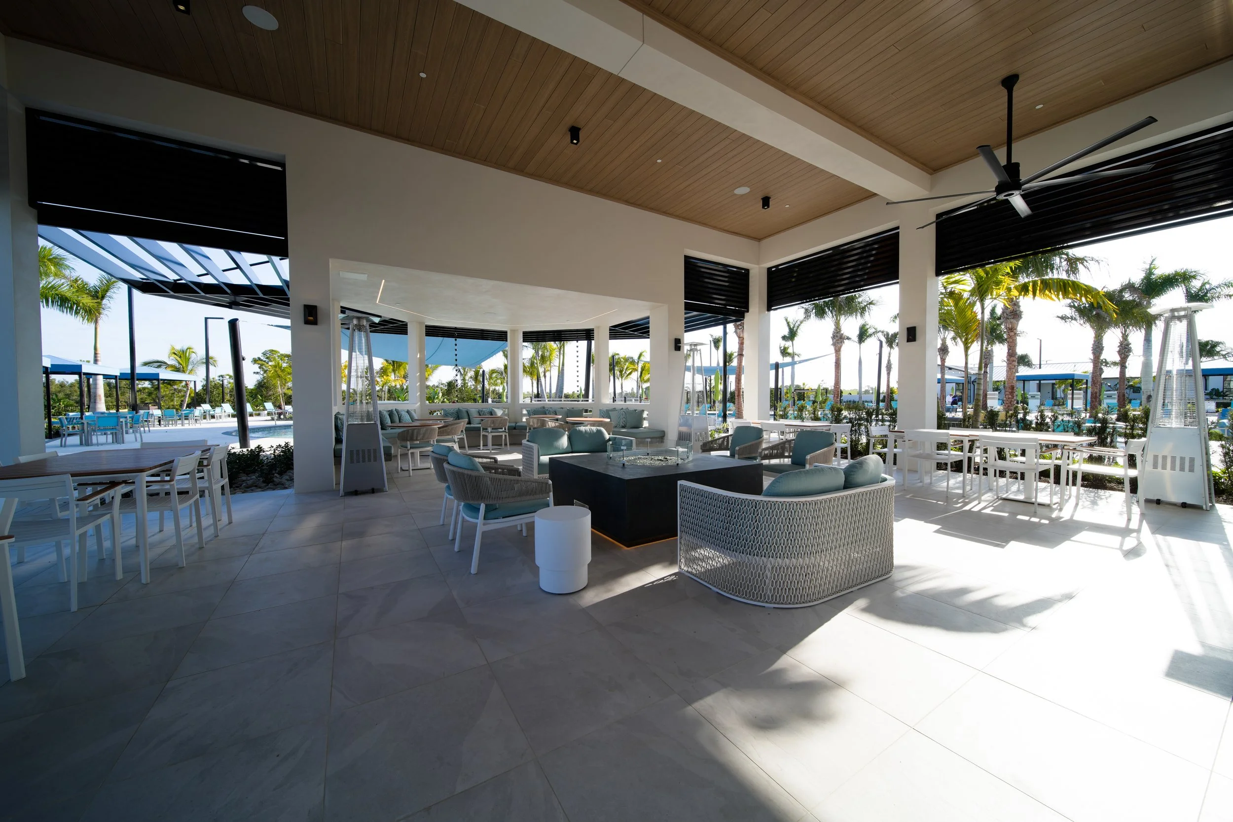 A modern outdoor lounge area with a wooden ceiling, white walls, and large windows. There are various lounge chairs, sofas, and tables. Outside, palm trees can be seen, along with patio dining tables and an outdoor pool area.