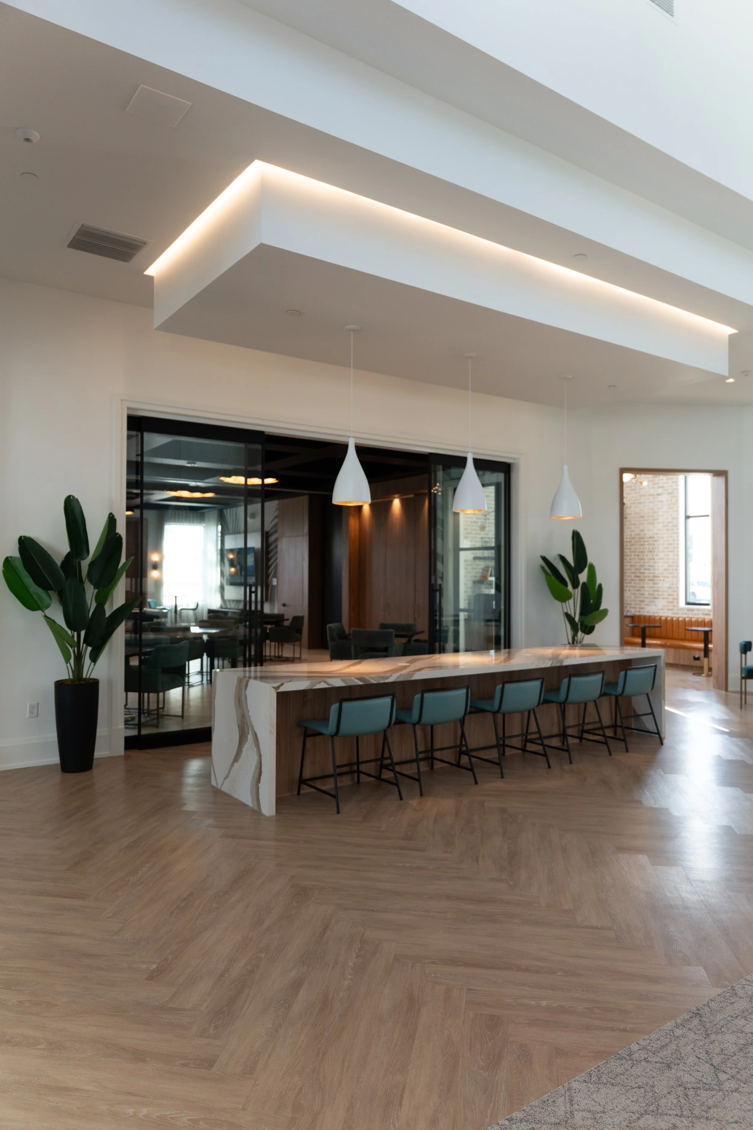 Modern hotel or restaurant interior featuring a large marble-top bar with blue-tufted chairs, white pendant lights, indoor plants, and a glass-walled seating area.
