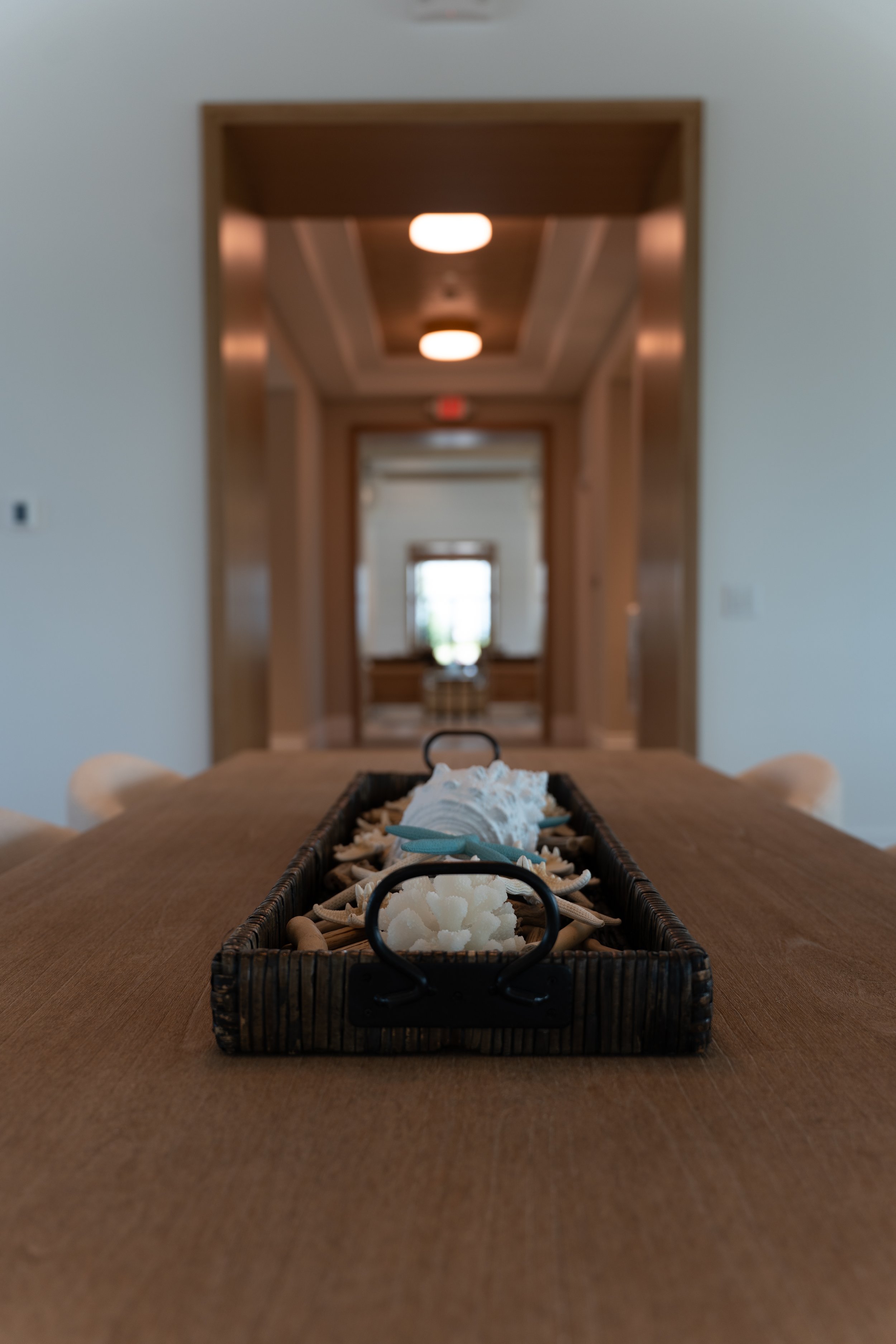 A rectangular basket filled with seashells and decorative white coral placed on a wooden table, with a blurred background of an interior hallway leading to a room.