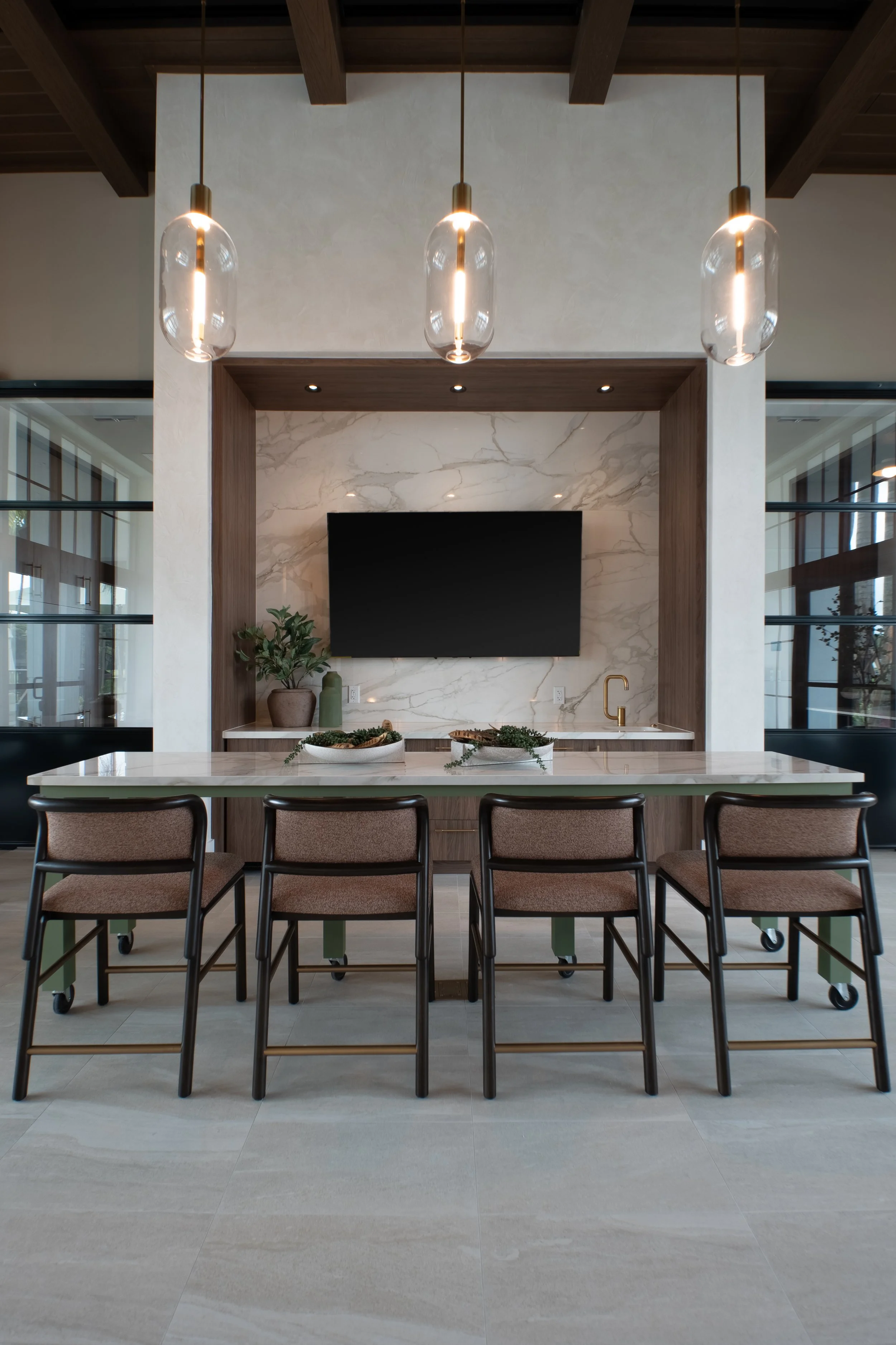 Modern kitchen with a large white marble island, six brown chairs, pendant lights, TV, and decorative plants.