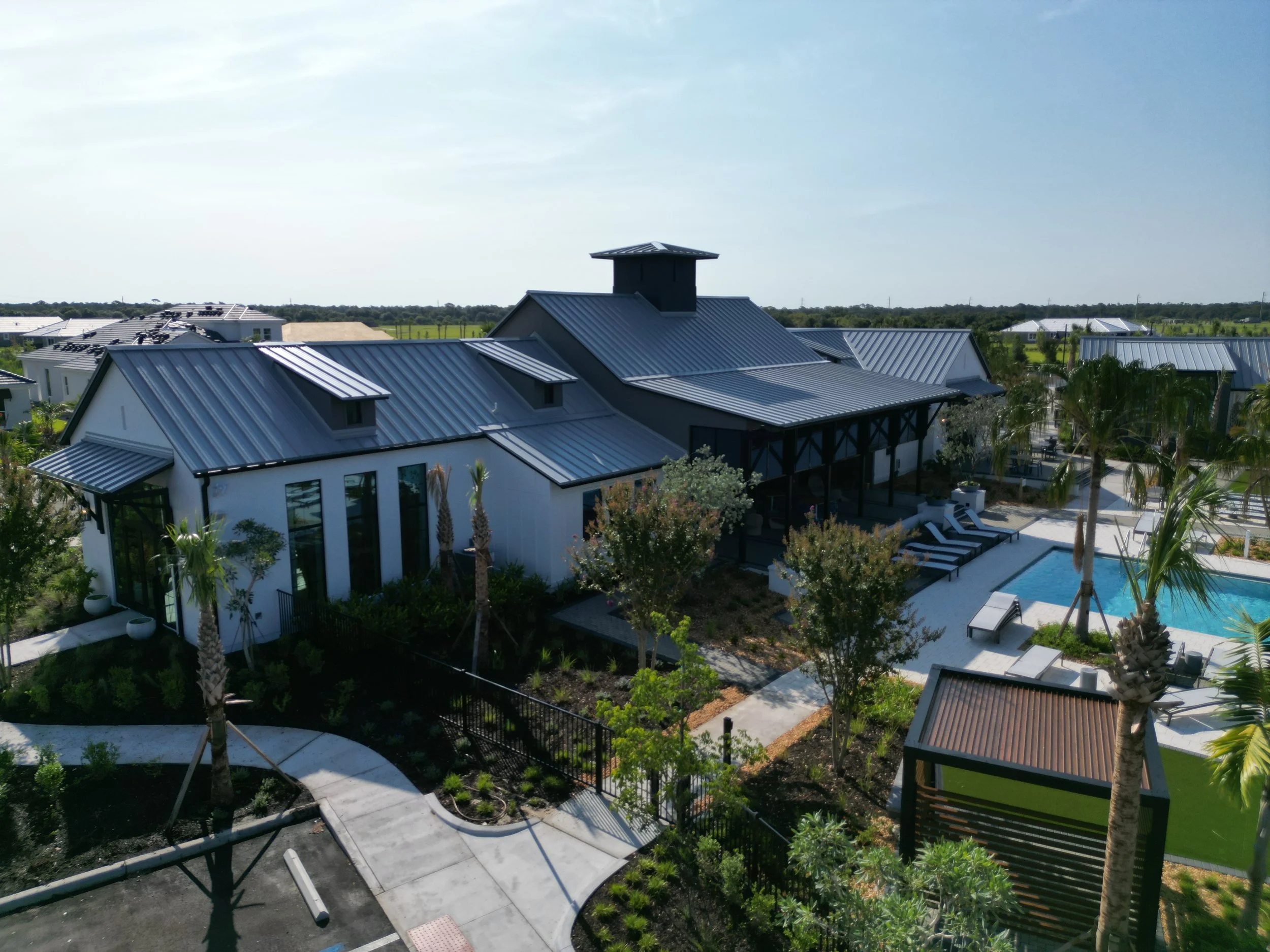 Aerial view of modern houses with metal roofs, lush greenery, a swimming pool, and a landscaped yard against a clear blue sky.