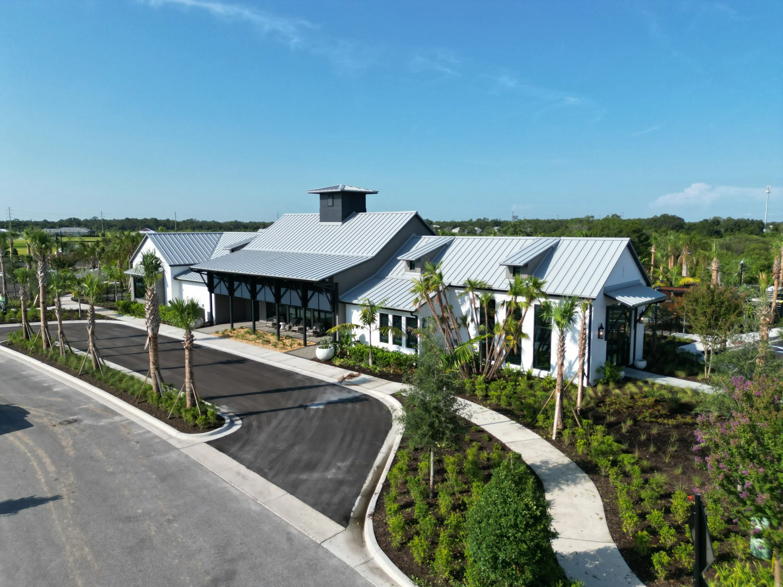 A modern building with white walls and a metal roof, surrounded by landscaped greenery including palm trees and flowering plants, under a clear blue sky.