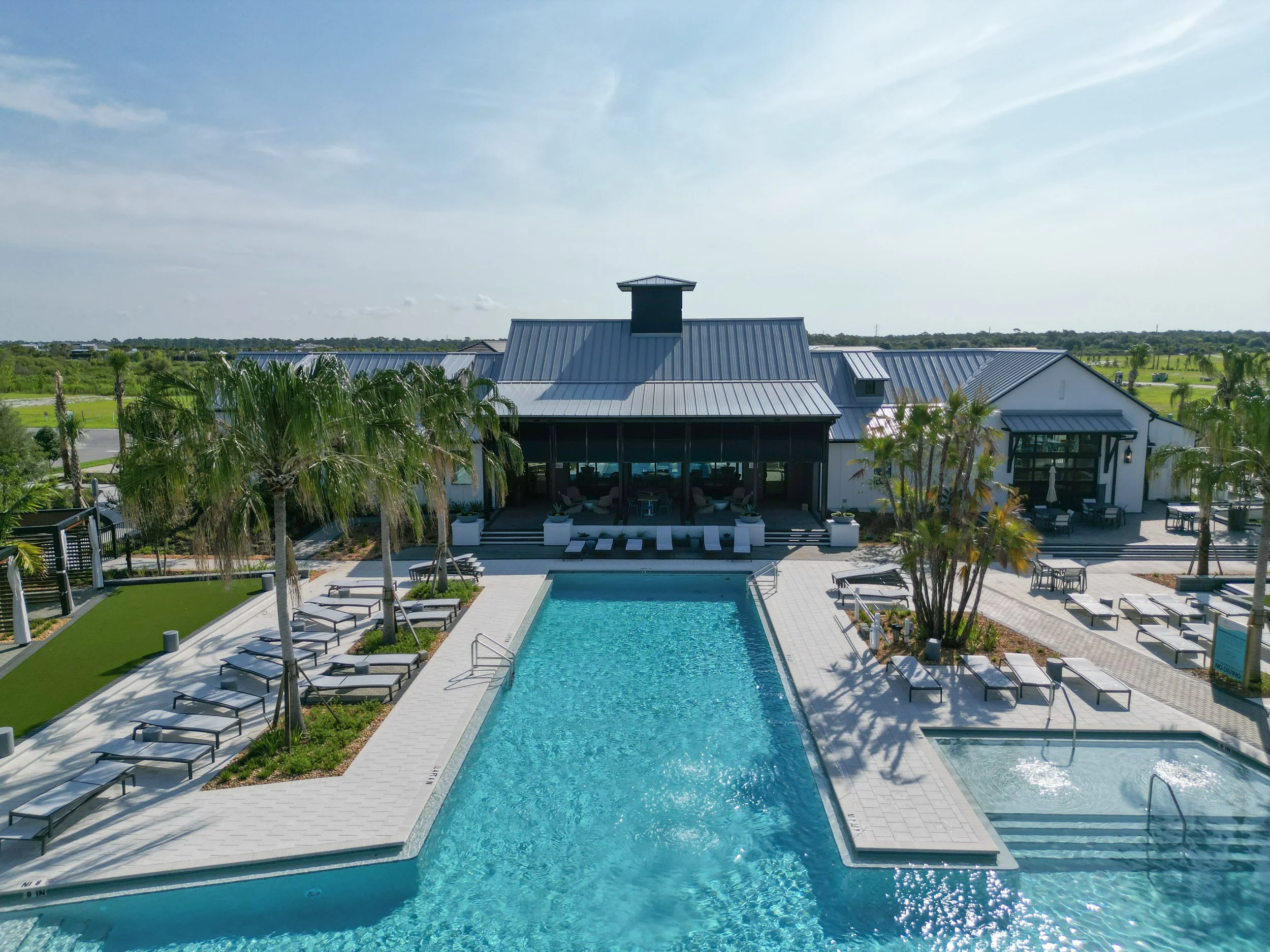 An outdoor swimming pool with a hot tub, surrounded by lounge chairs and palm trees, with a modern building and green landscape in the background under a partly cloudy sky.