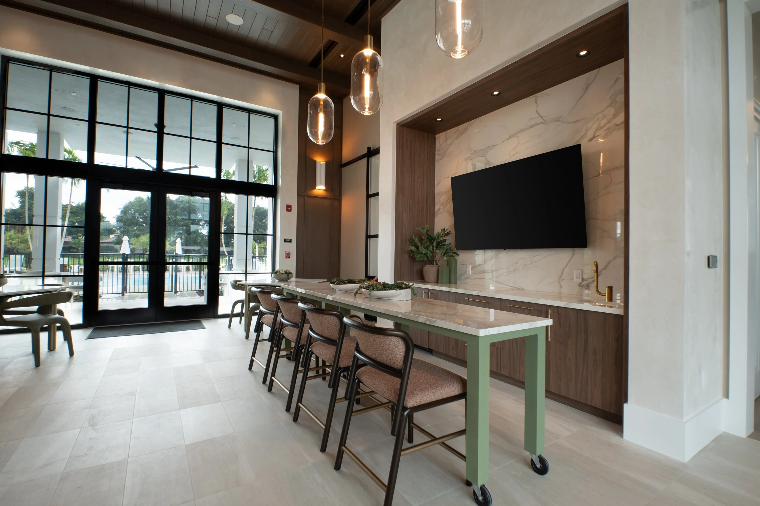 Modern indoor space with large windows, hanging pendant lights, a marble wall with a mounted flat-screen TV, and a table with chairs and potted plants.