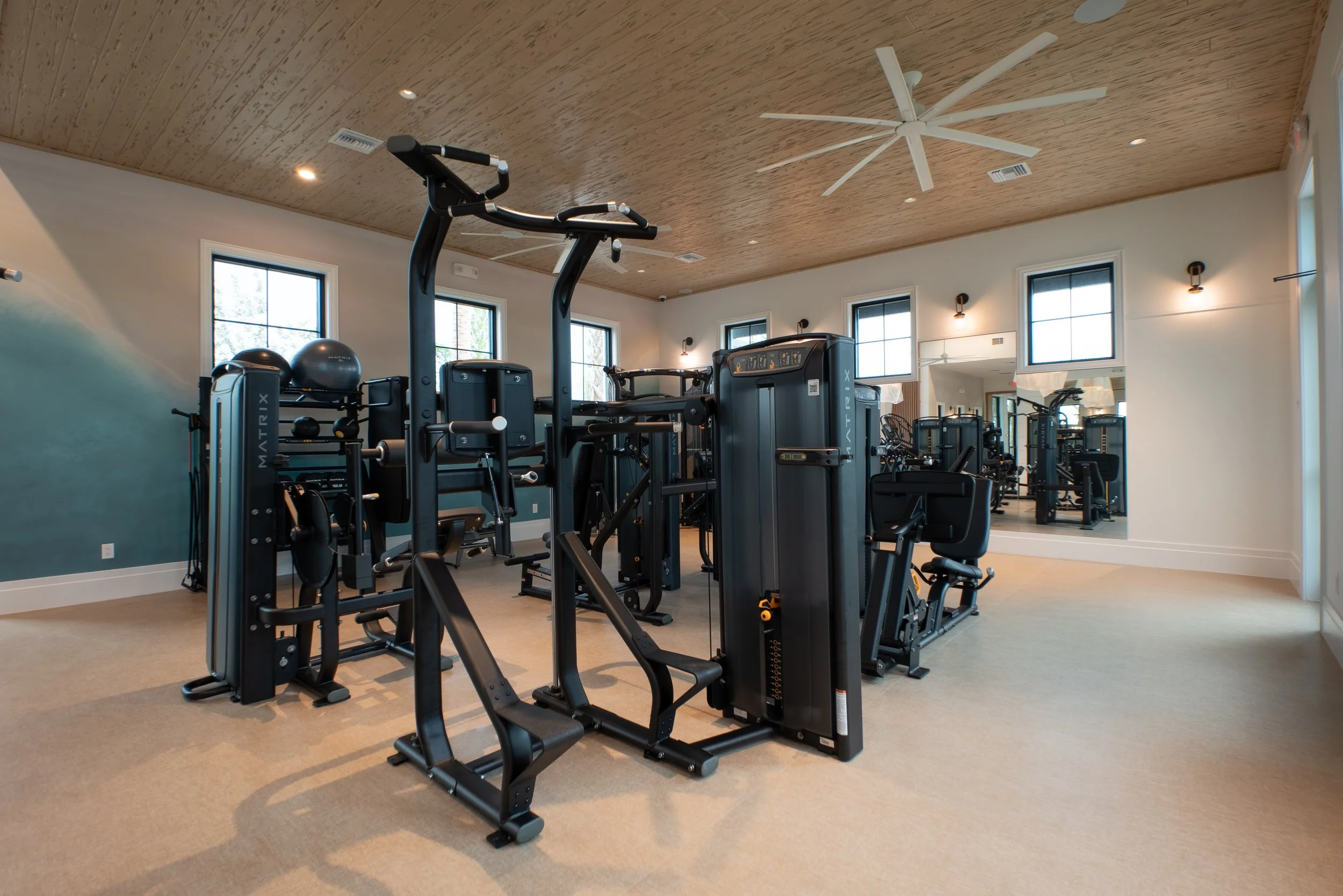 A fitness room with various exercise equipment including cardio machines and weights, with large windows, a wooden ceiling, and modern lighting.