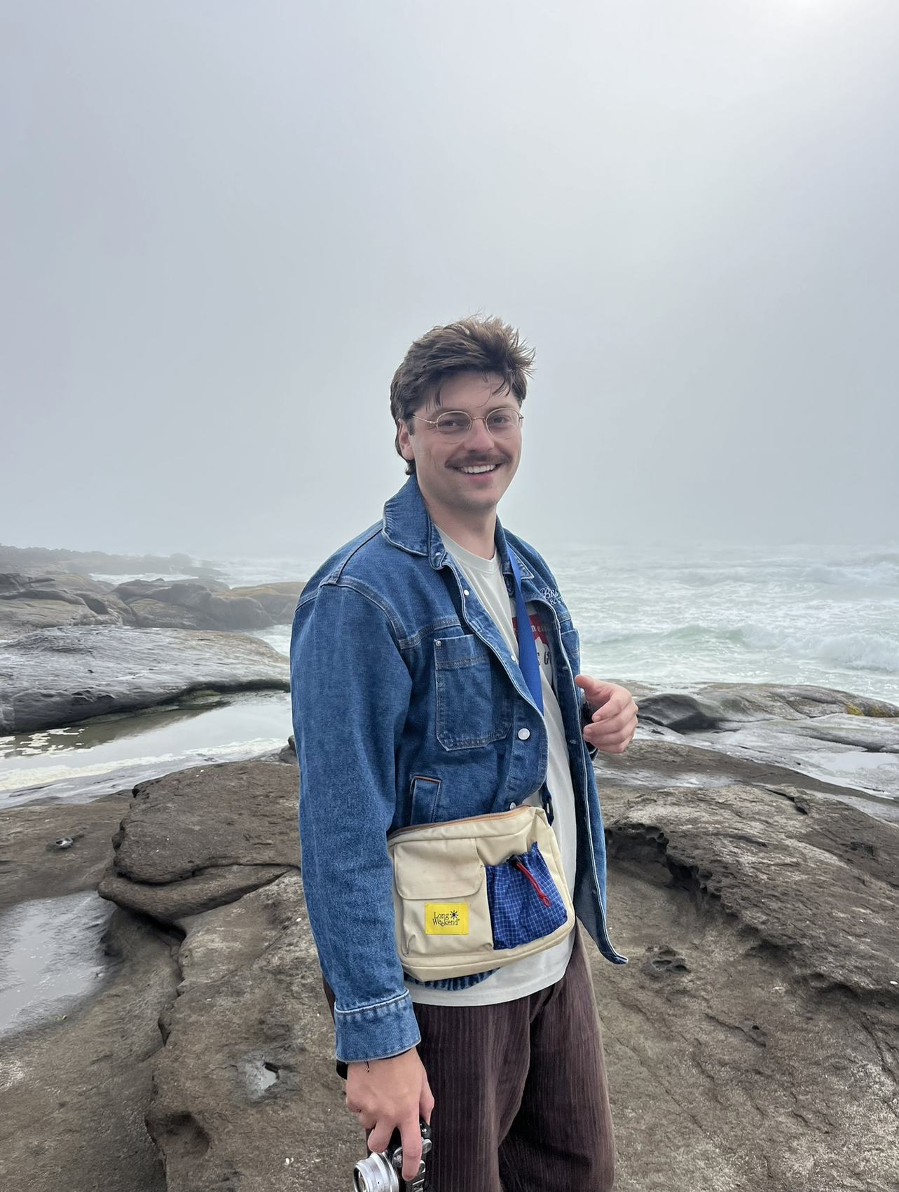 A man wearing glasses and a denim jacket stands on a rocky beach with ocean waves and overcast sky in the background, smiling at the camera.