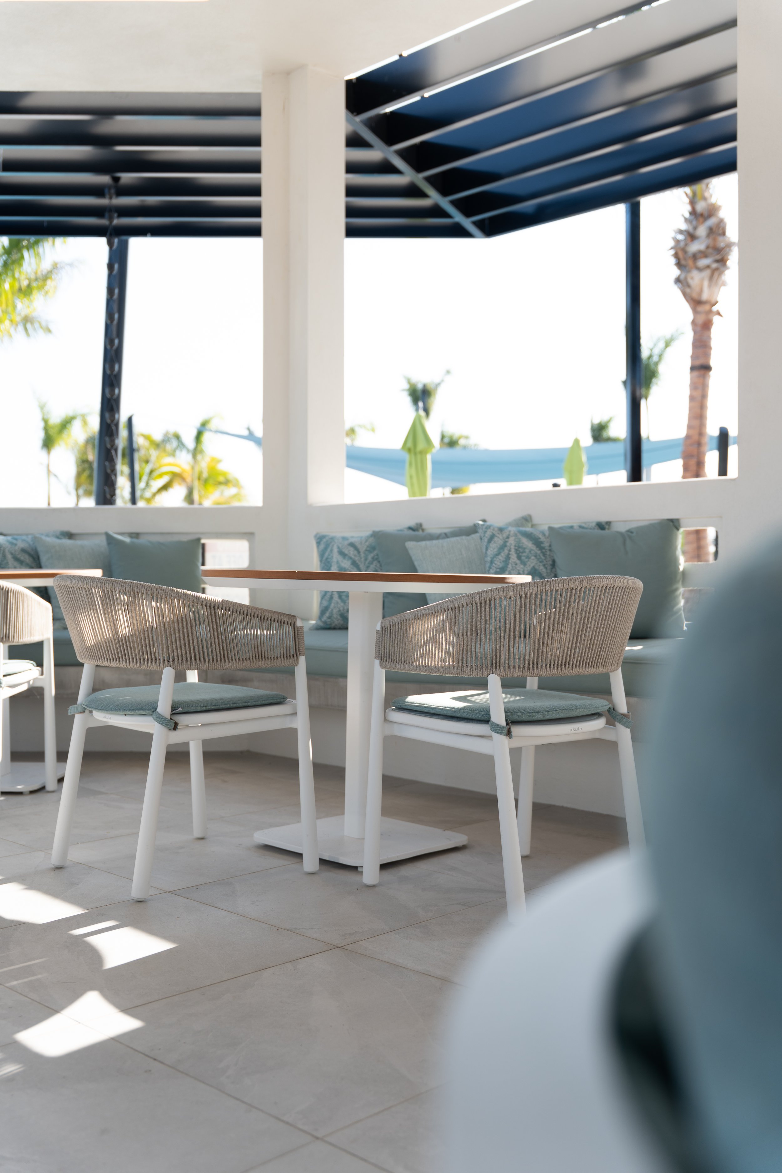 Outdoor seating area with white chairs and a table, cushions on the bench, with a shaded patio and palm trees in the background.