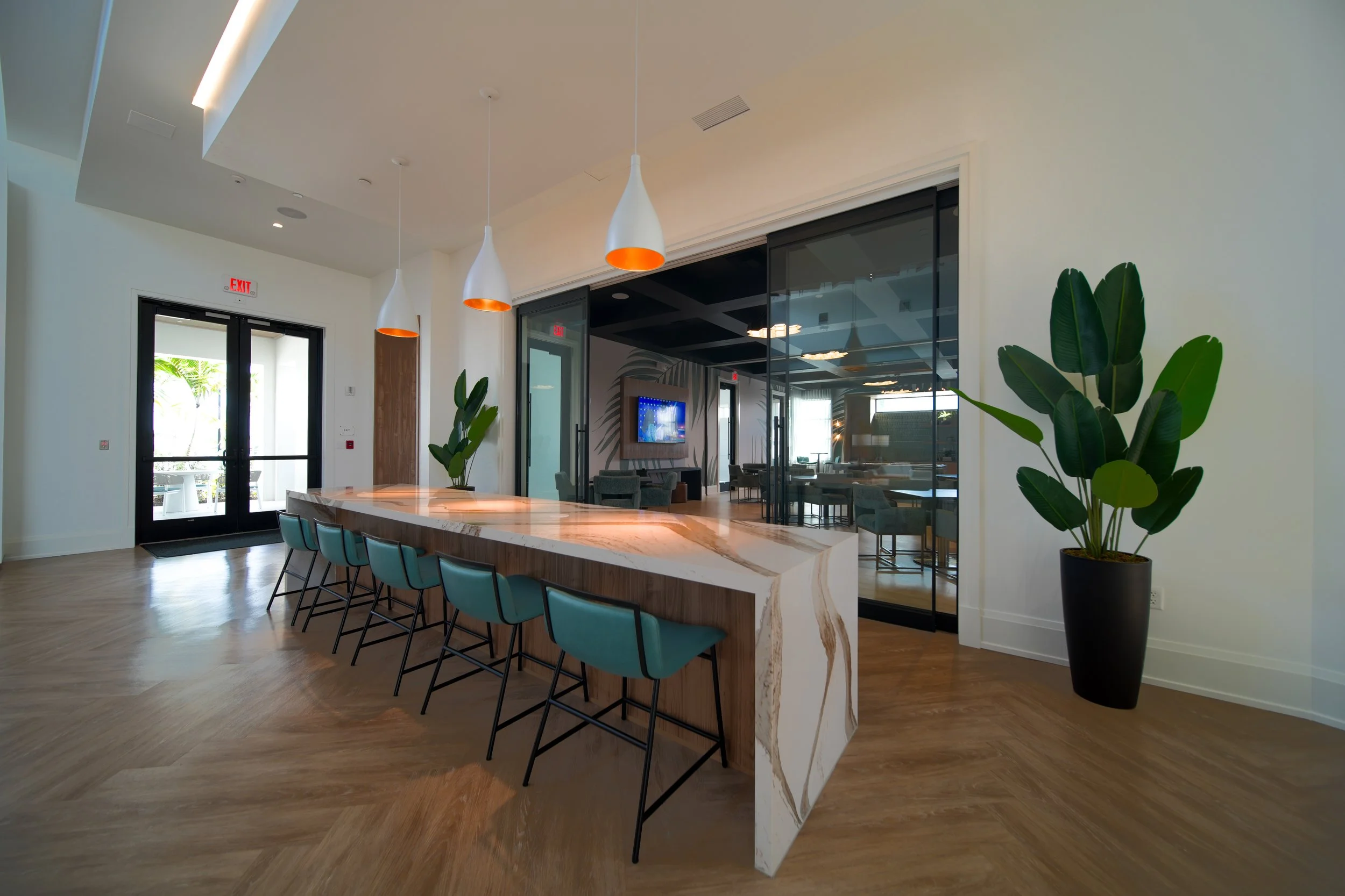 Modern interior space with a marble-topped central island, teal chairs, hanging white pendant lights with orange interior, large potted plant, glass partitions, and a television in the background.