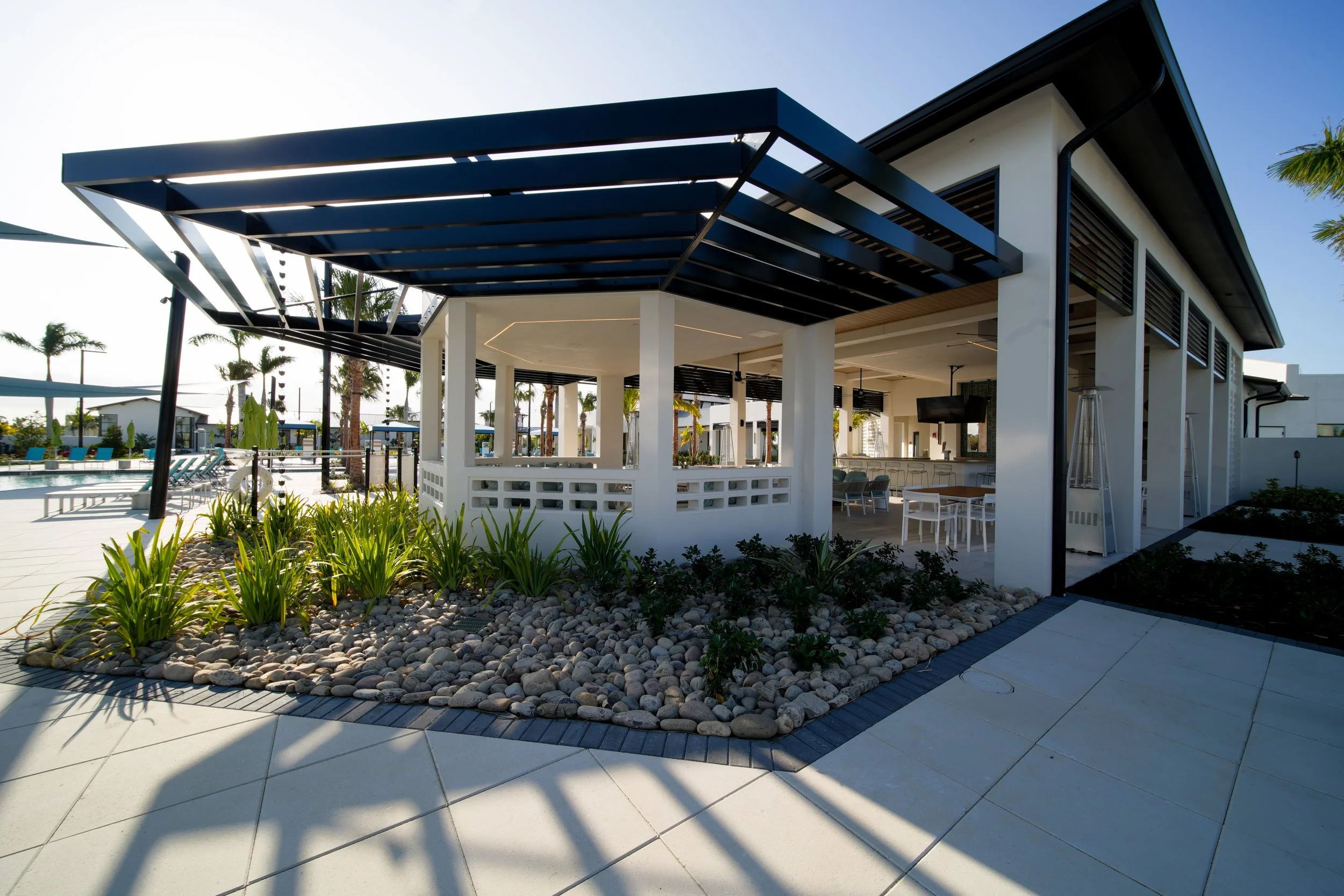 Modern building with black overhang, outdoor seating area, palm trees in background, and landscaped garden with rocks in foreground.