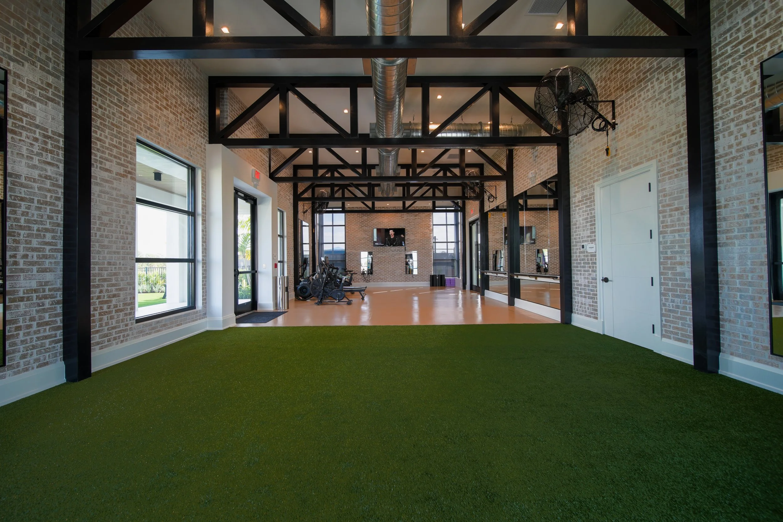 A modern gym with brick walls, large windows, black metal beams, ventilation duct, ceiling fan, exercise bikes, and a wall-mounted television, with artificial turf flooring.