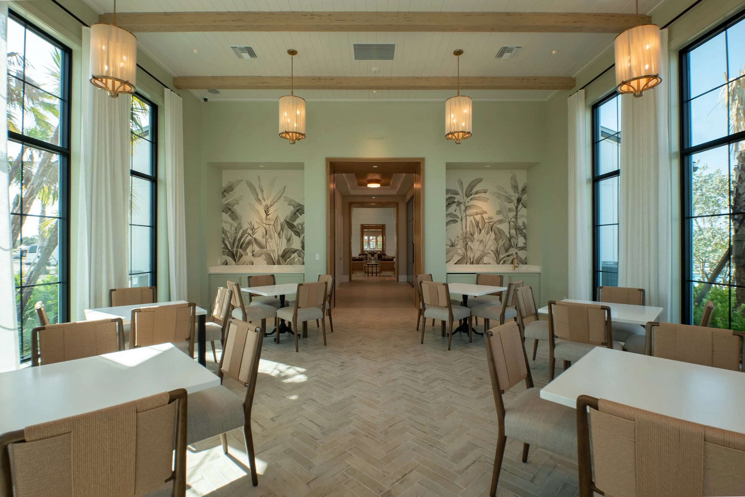 Interior of a bright dining area with large windows, beige chairs, white tables, and wall art with botanical designs.