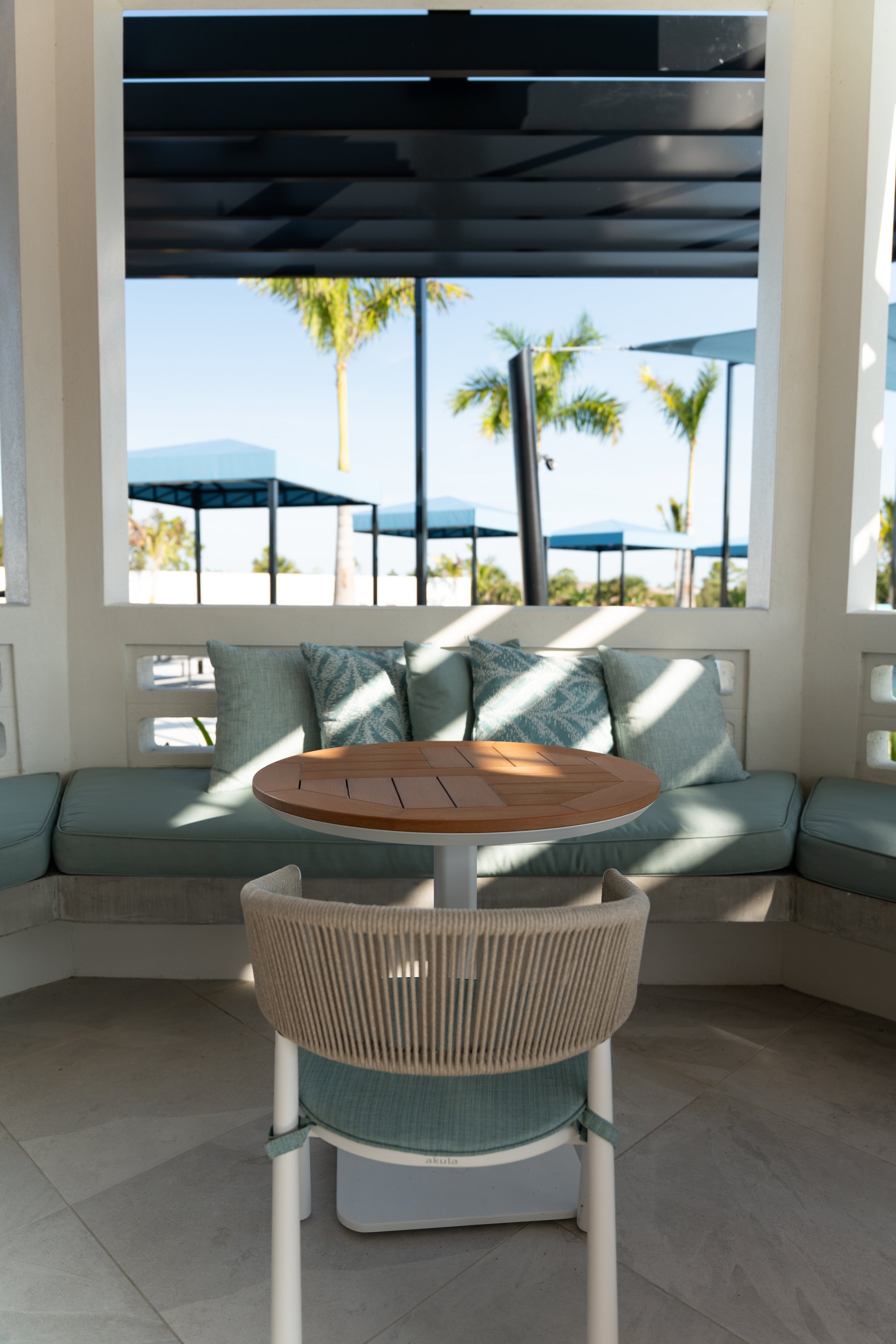 Outdoor patio area with a cushioned sofa, a round wooden table, a wicker chair, and a view of palm trees and blue umbrellas in the background.