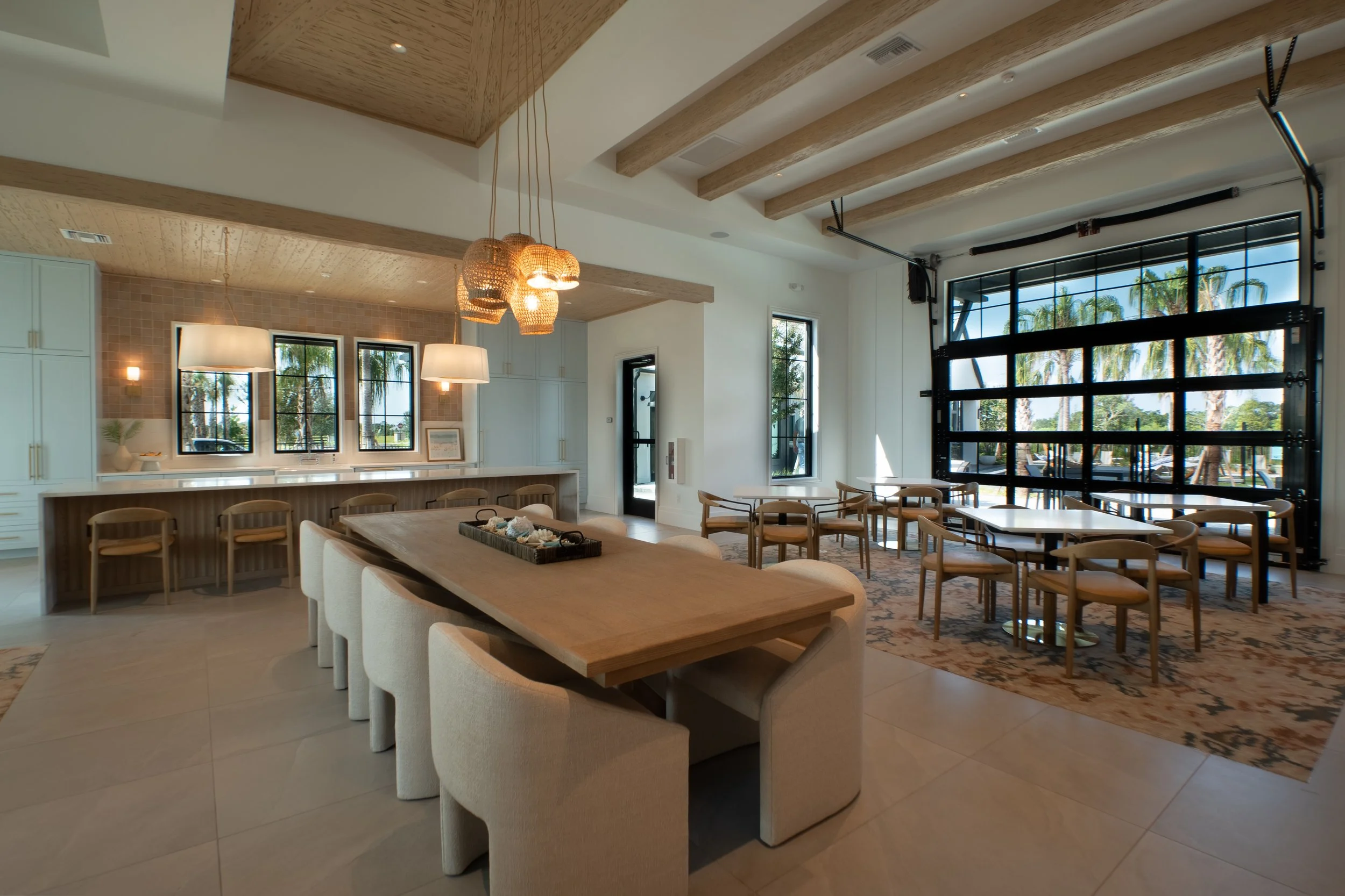 Interior of a modern dining area with a long wooden table surrounded by cream-colored upholstered chairs, light gray cabinets, pendant lights, large windows with views of palm trees, and a decorative rug.