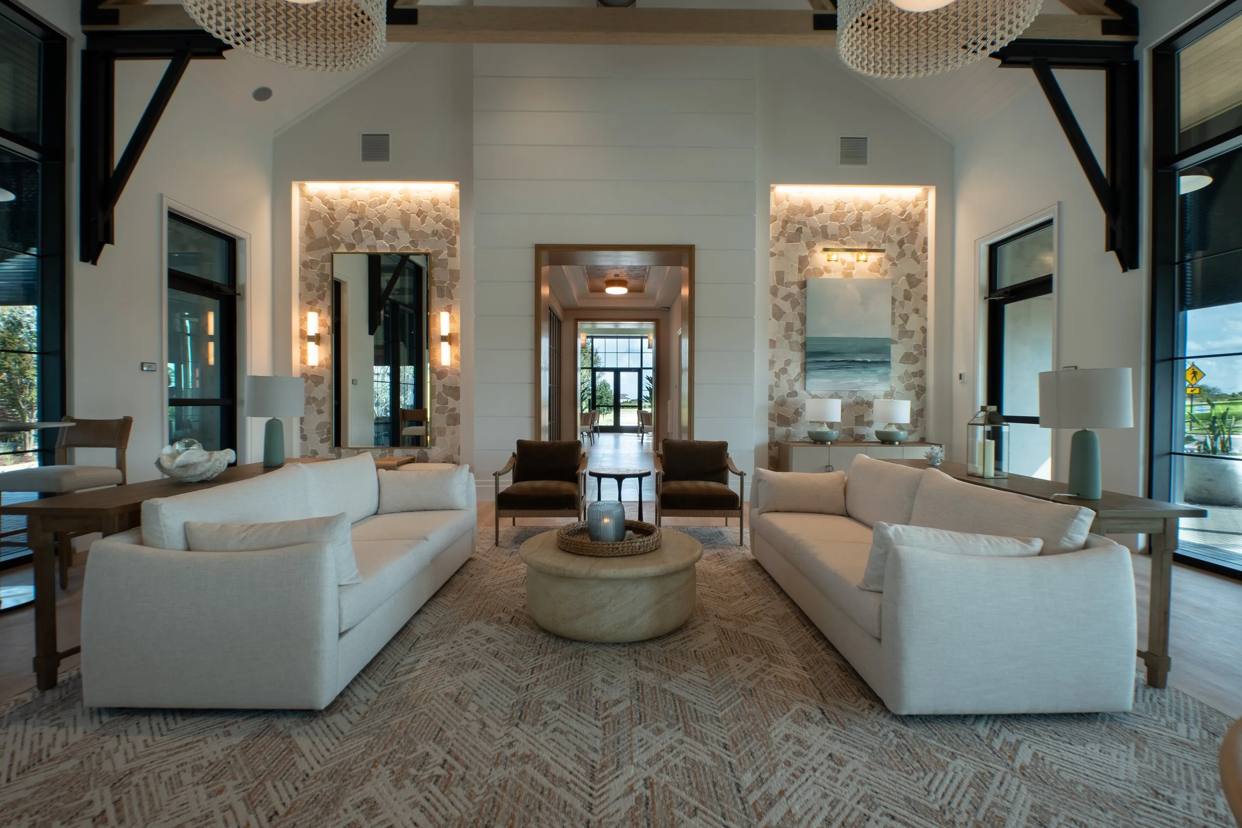 Interior of a modern living room with white sofas, a round coffee table, and a patterned rug. Large windows let in natural light. The walls are decorated with art and mirrors, with lighting fixtures and lamps providing additional illumination.