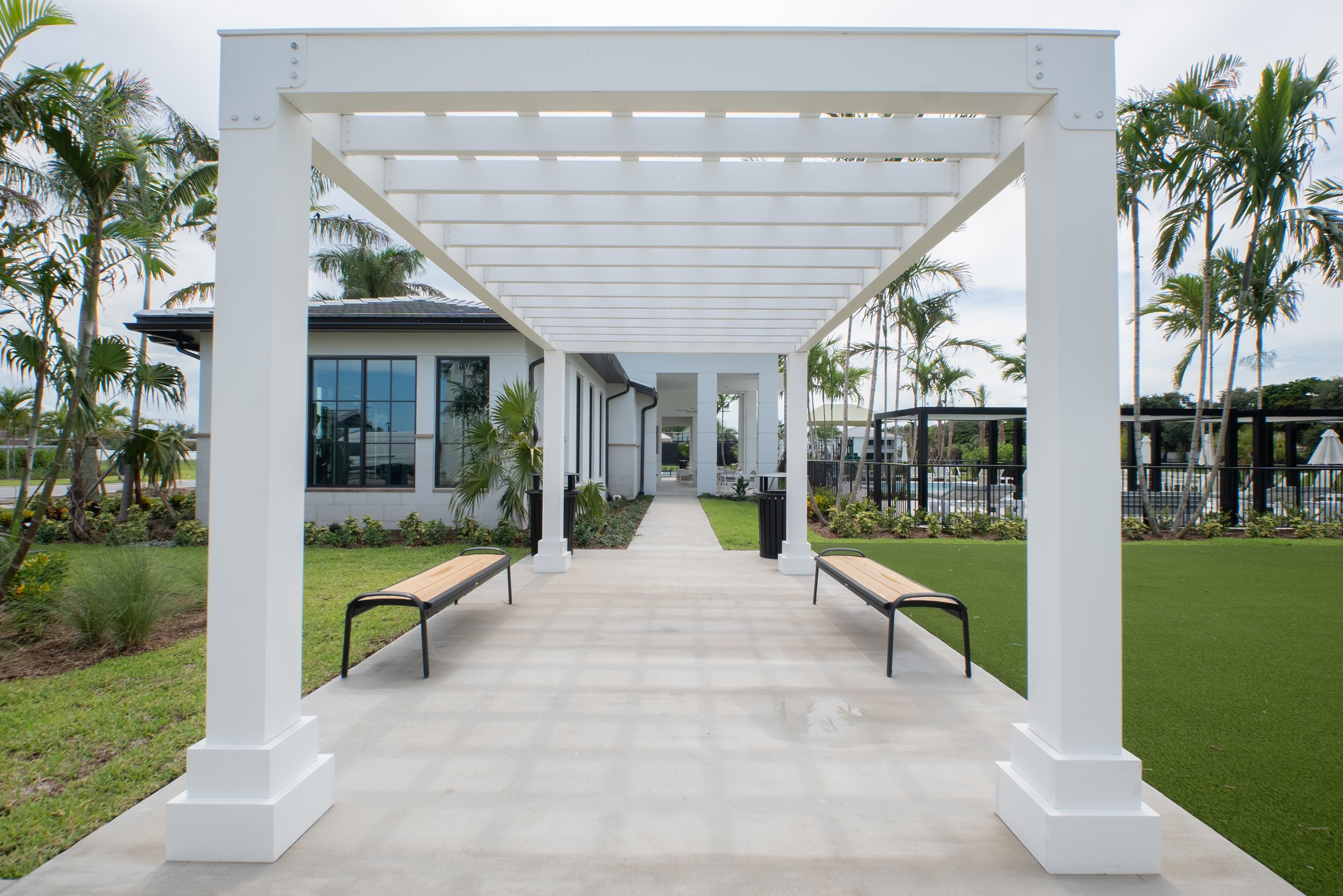 Outdoor patio with white pergola, black benches, and surrounding greenery including palm trees at a modern residential or commercial building.