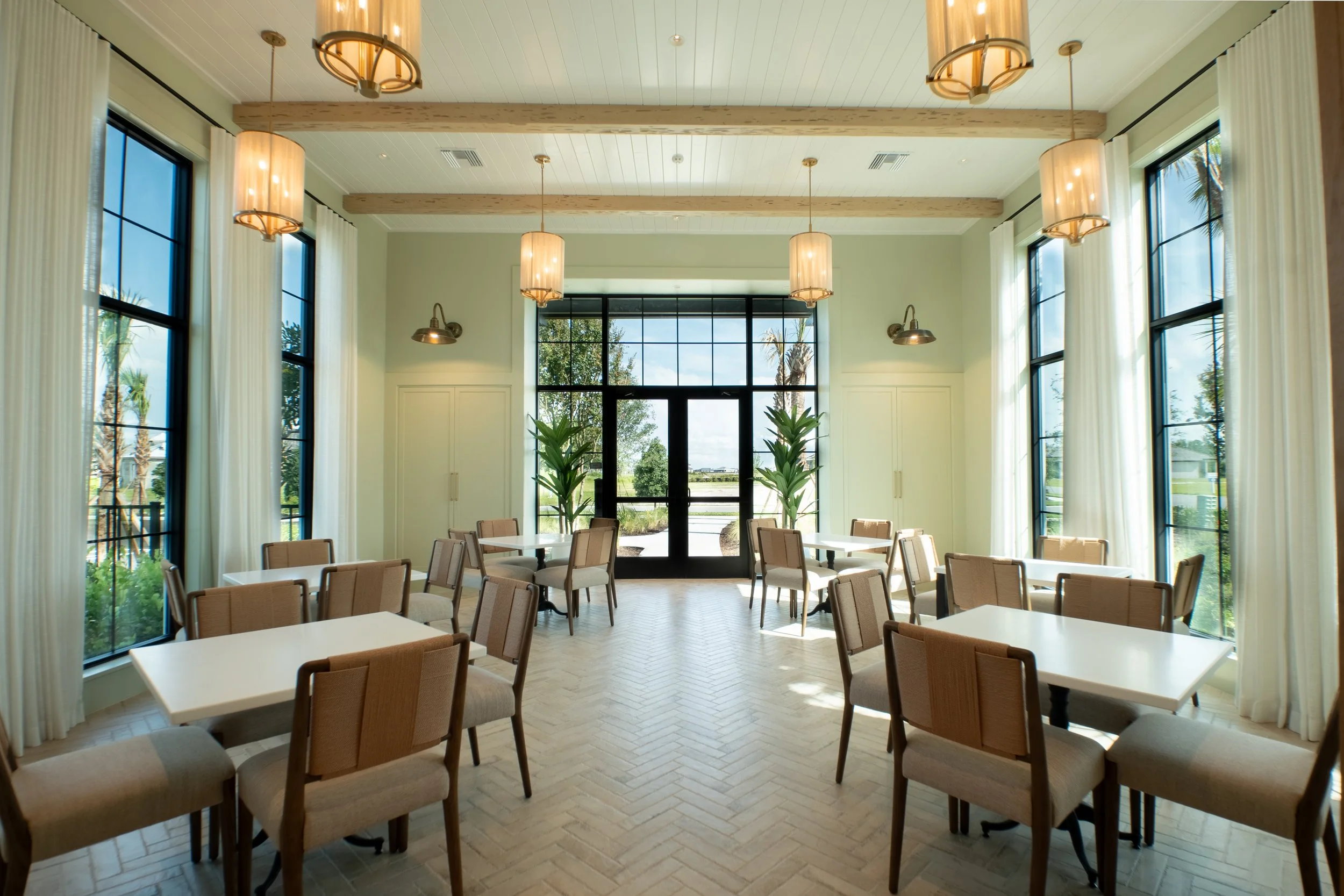 Sunlit dining room with large windows, beige chairs around white tables, ceiling lights, and plants near the entrance door.