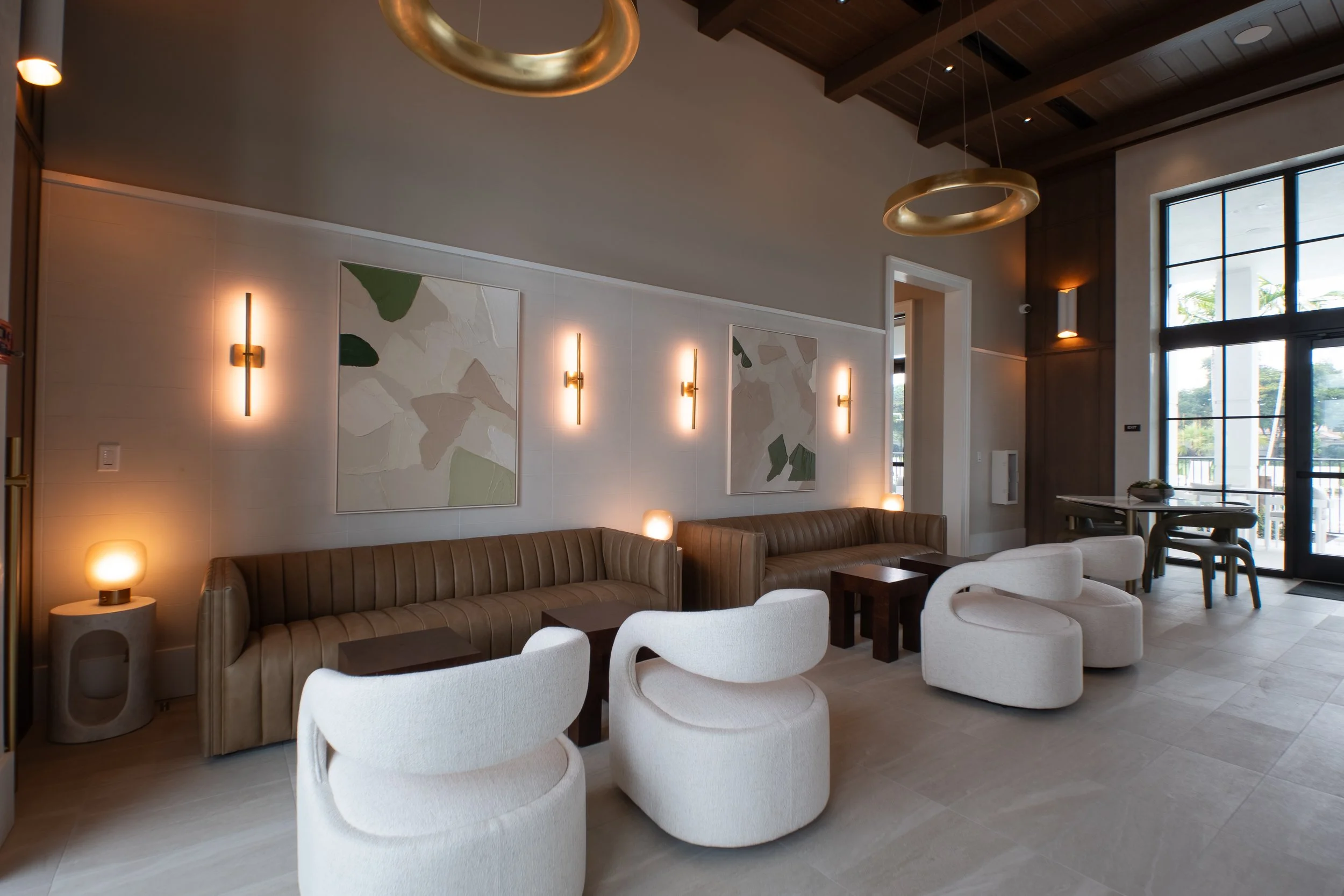 A modern, stylish indoor lounge area with beige and white seating, abstract art on the wall, warm lighting, and large windows letting in natural light.