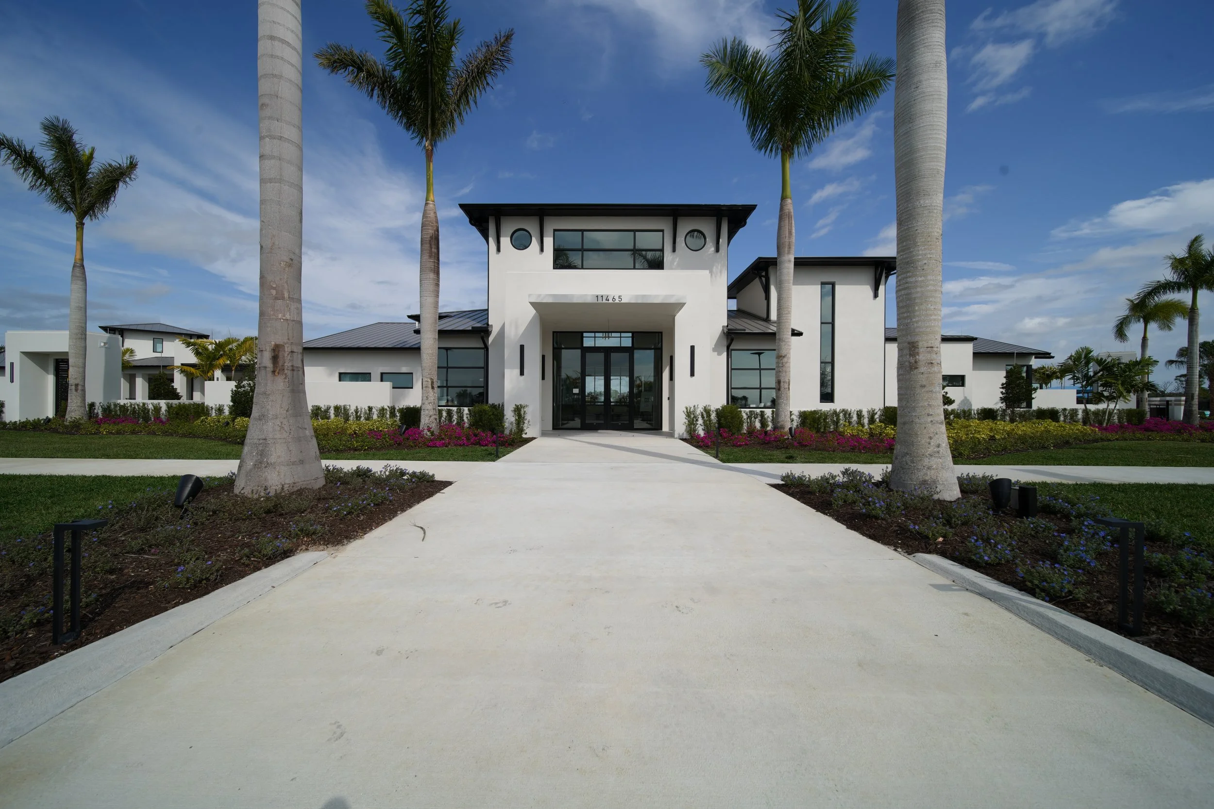 Modern white house with large glass windows, surrounded by palm trees and landscaped garden, with a concrete walkway leading to the entrance.